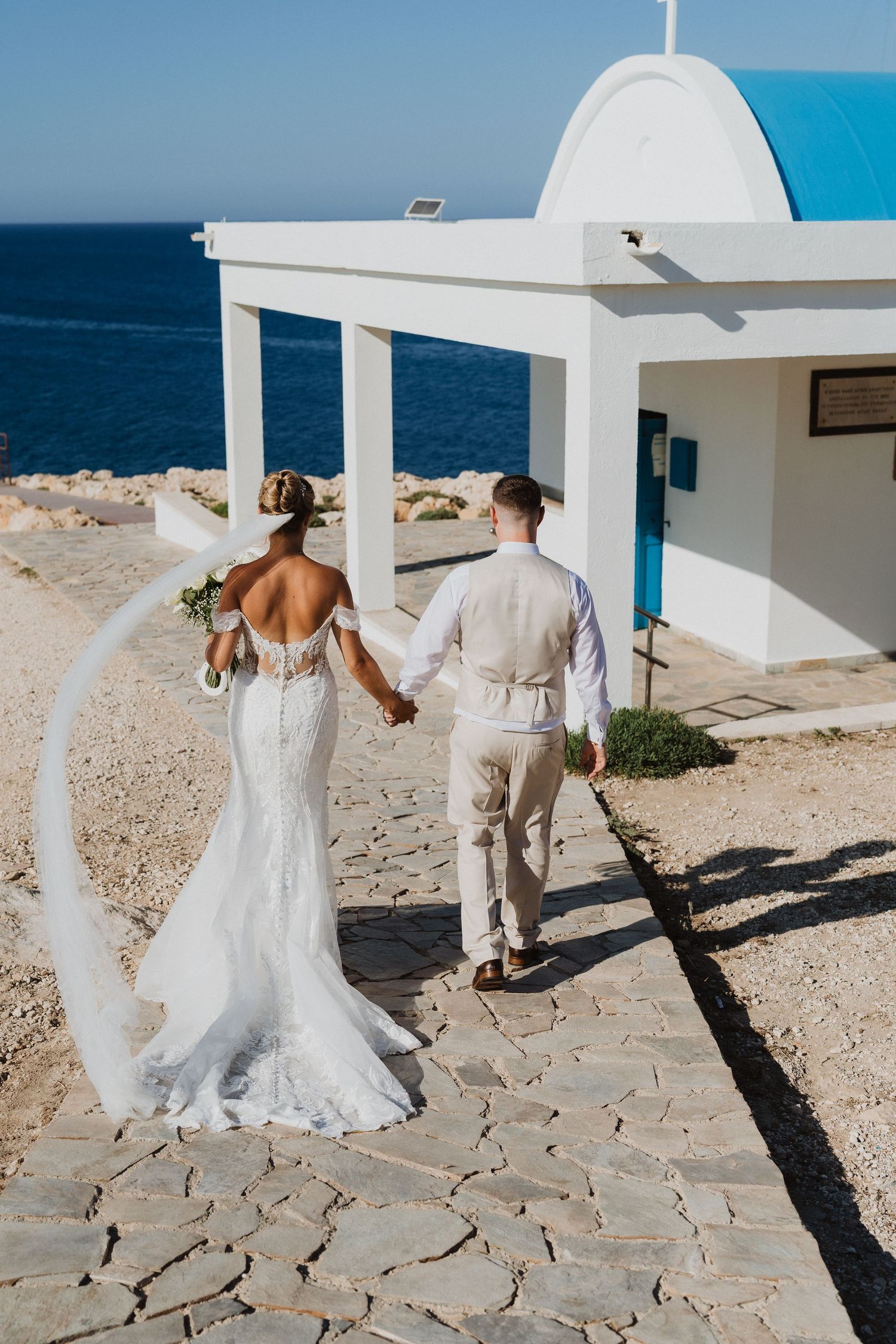A couple in wedding attire dances playfully on a Mediterranean seaside terrace with white walls and ocean views.