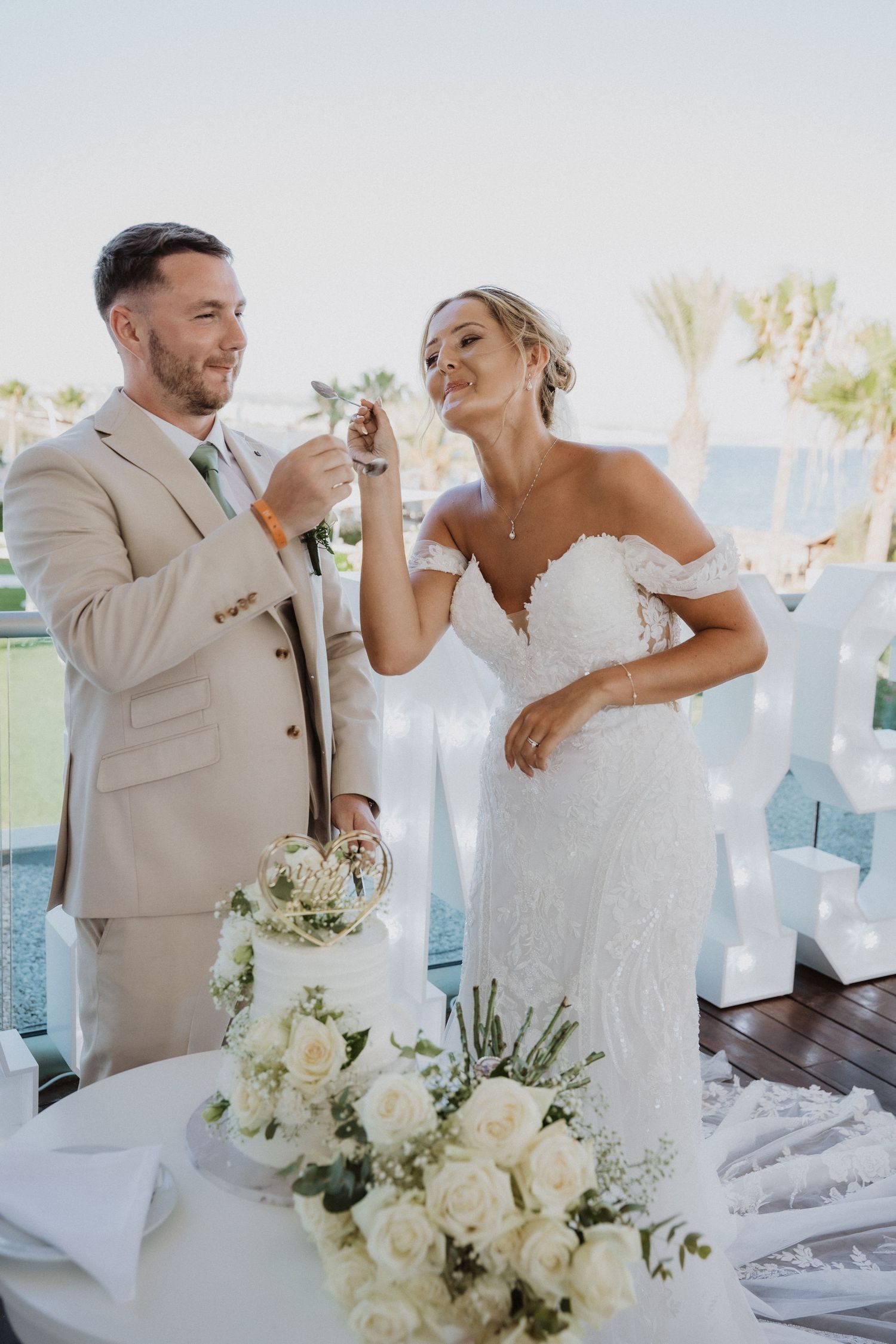 A couple in wedding attire shares a playful moment during their reception, surrounded by white floral arrangements.