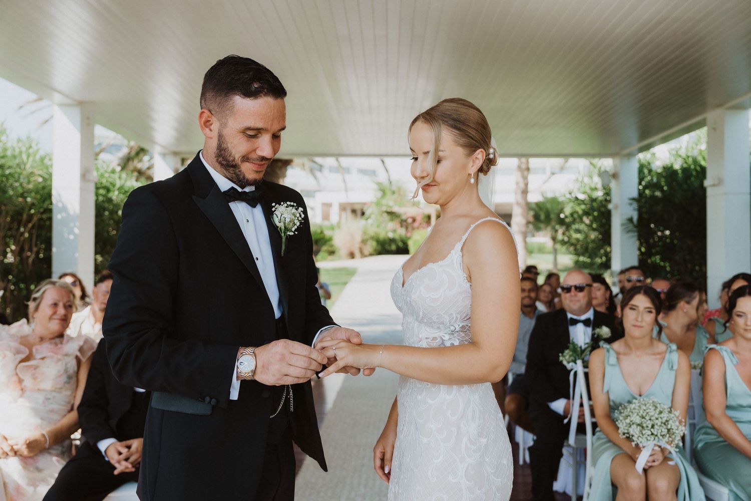 A couple exchanges rings during their wedding ceremony under a covered pavilion surrounded by guests.