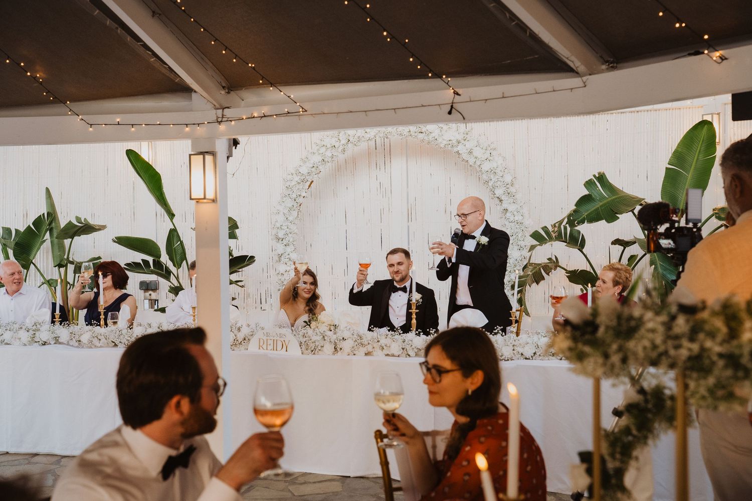 Wedding guests raise glasses during a toast at an elegant white floral reception venue.