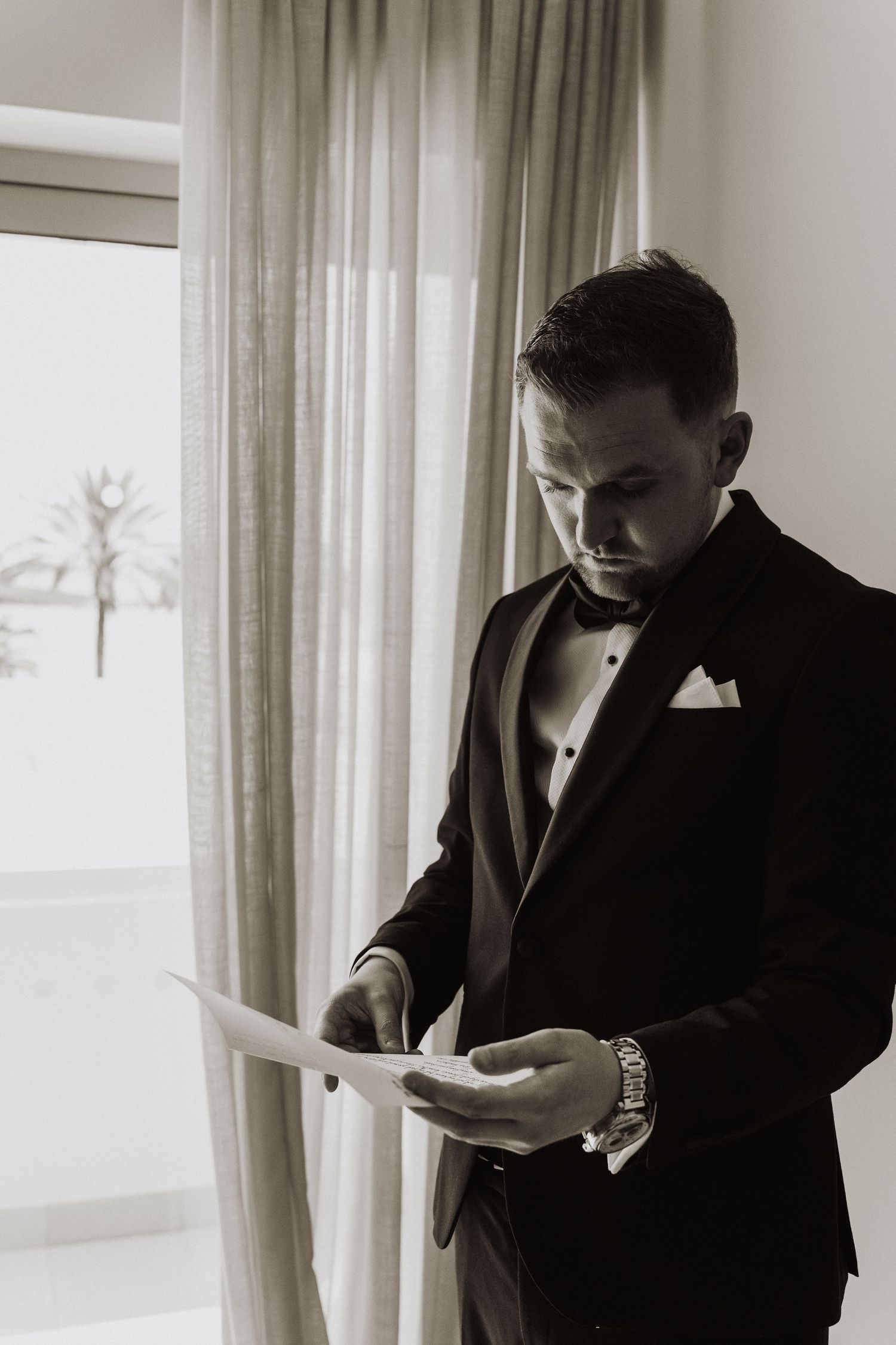 Groom in black tuxedo reading wedding vows letter by window in black and white photo.