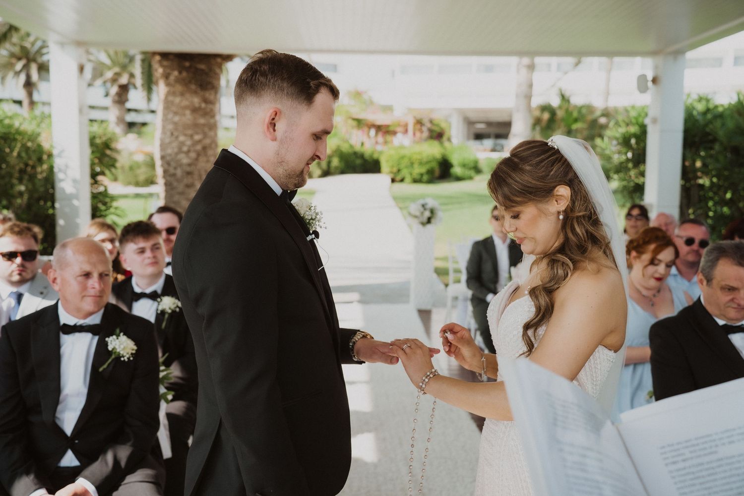 Bride placing wedding ring on grooms finger during outdoor ceremony under pavilion.