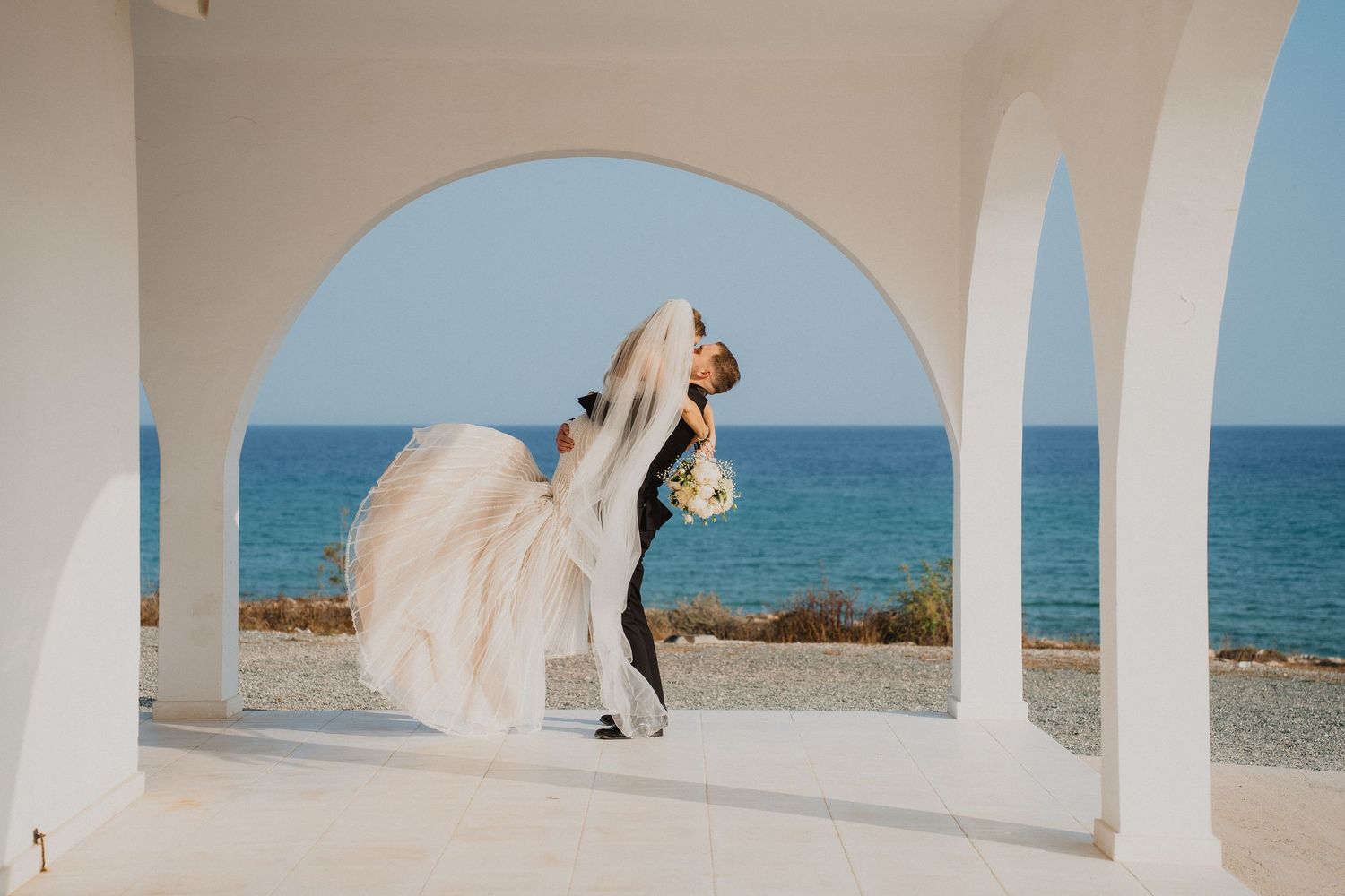 Groom lifts bride in flowing white gown under white arched columns with blue ocean views in background.