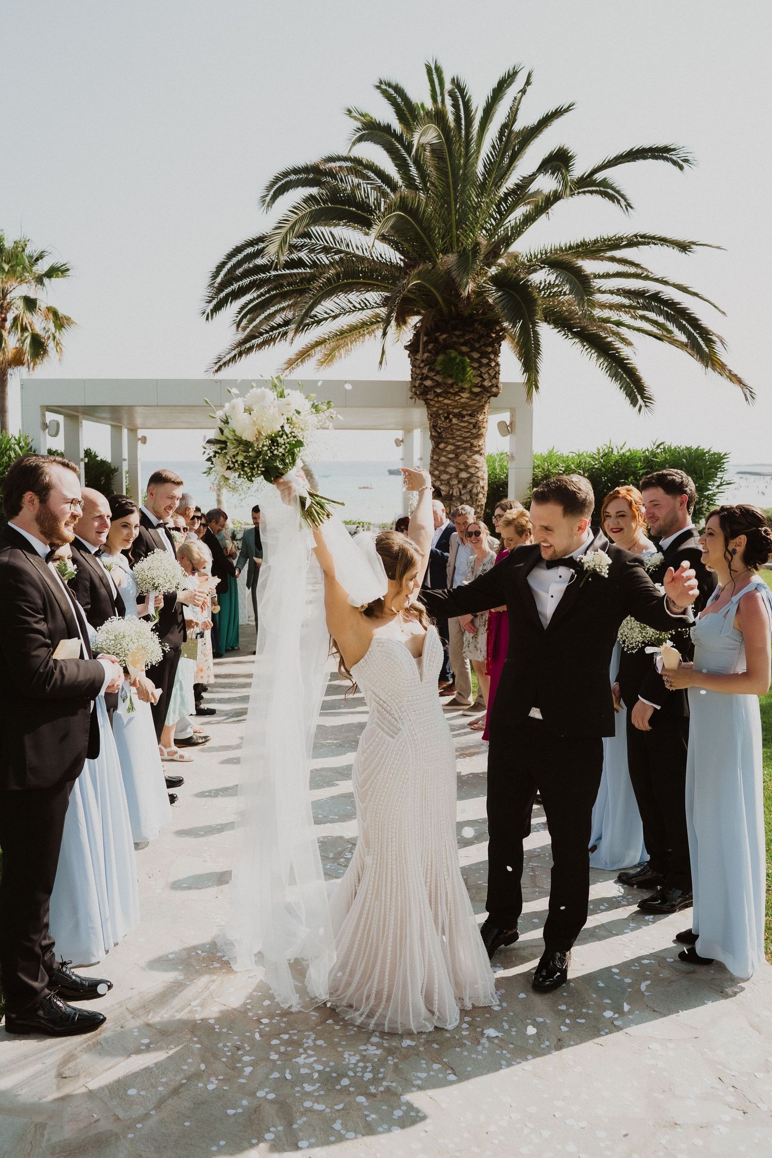 Bride raises bouquet joyfully as couple exits wedding ceremony past guests and palm trees.