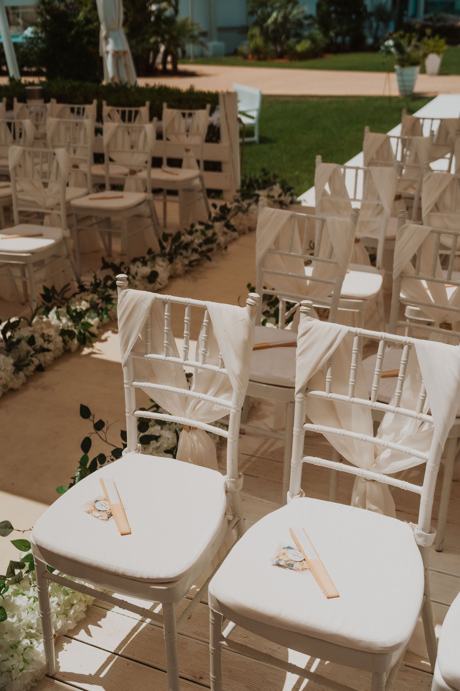 Elegant white Chiavari chairs with sheer fabric bows arranged for an outdoor wedding ceremony on a sunny day.
