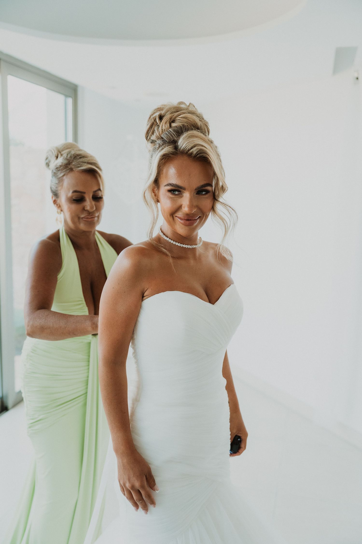 Bride in white strapless gown with pearl necklace smiles as bridesmaid in mint green dress helps fasten her wedding dress.