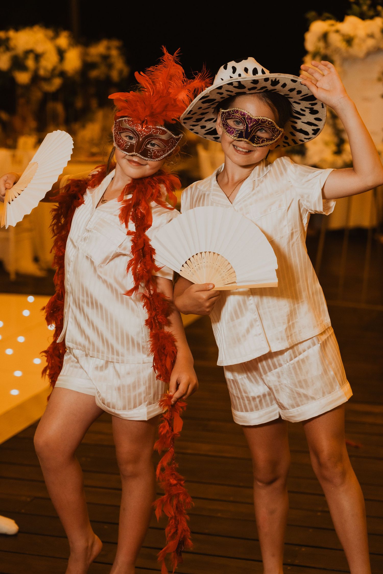 Two girls in white pajamas wear masquerade masks, feather boa, and hat while holding decorative fans at a party.