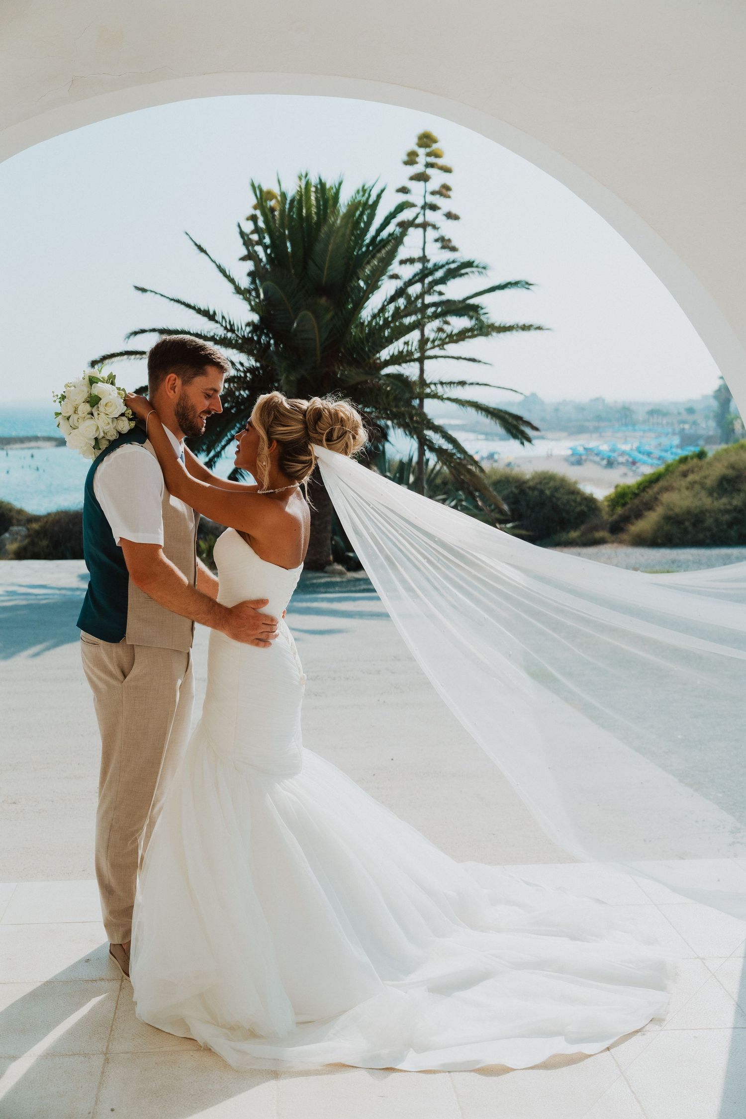 Bride and groom embrace under white arch with ocean view, veil flowing in breeze, palm trees in background.