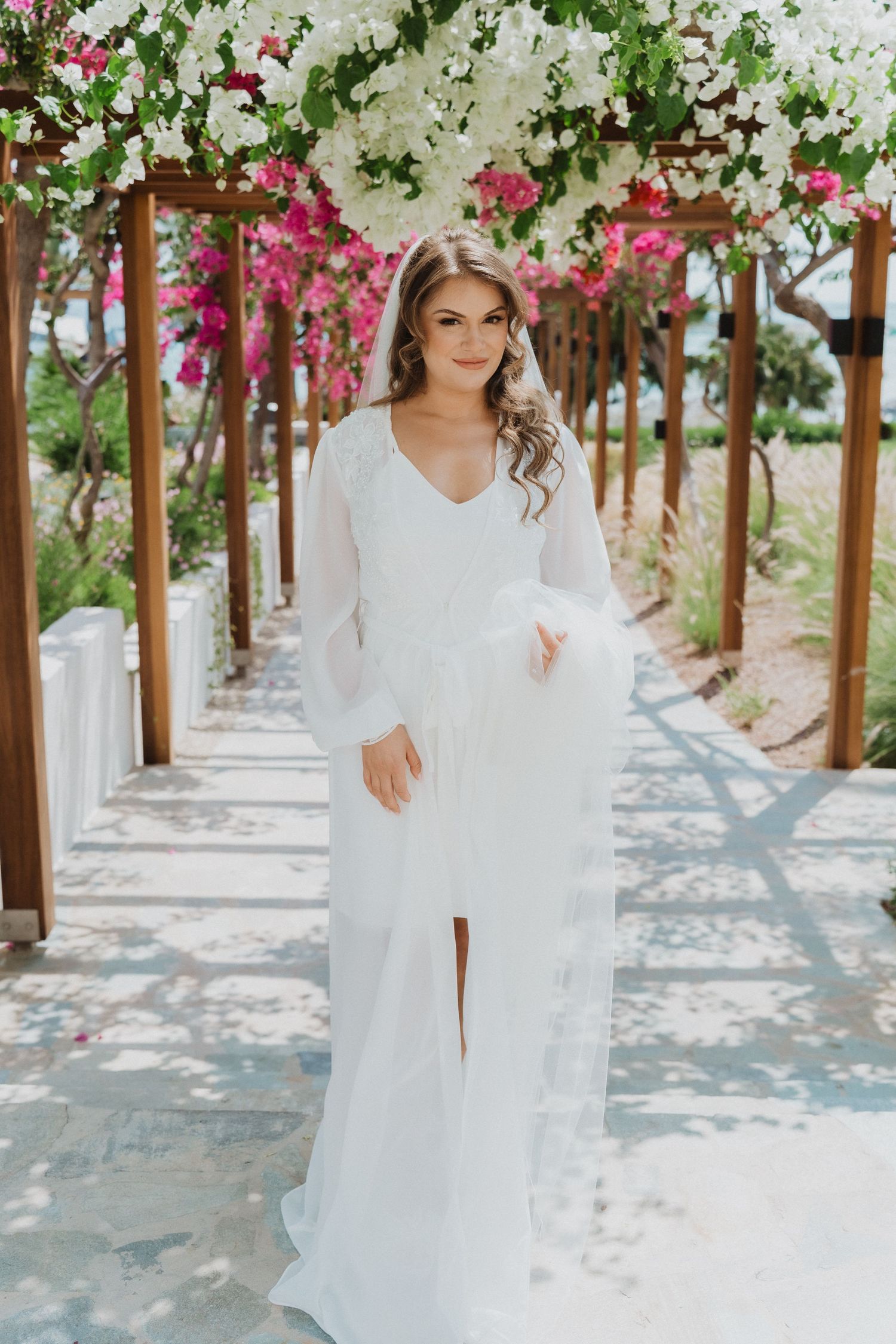 Bride in flowing white sheer robe standing under floral pergola arch.