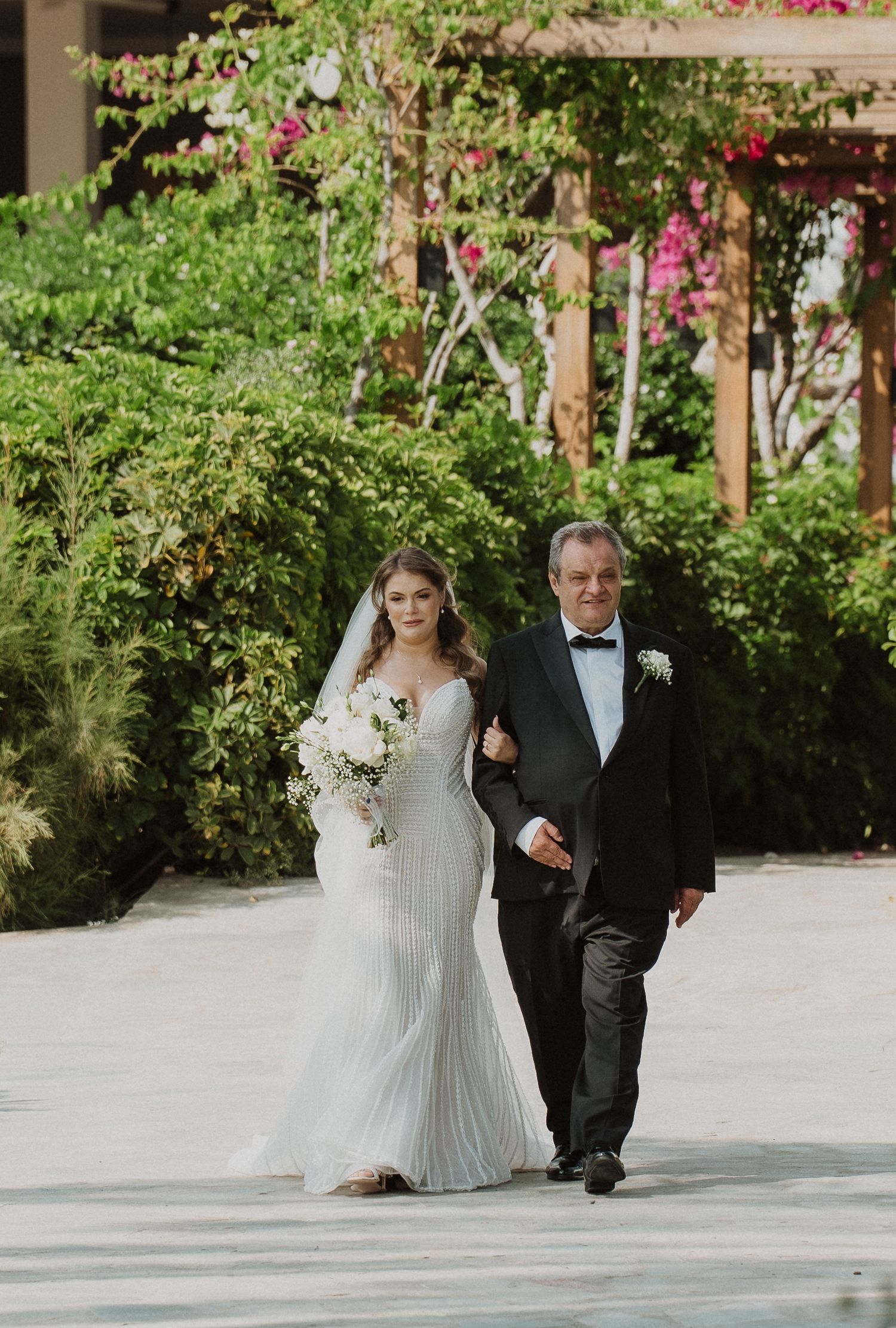 Bride in beaded gown walking down garden path with father in black tuxedo on wedding day.