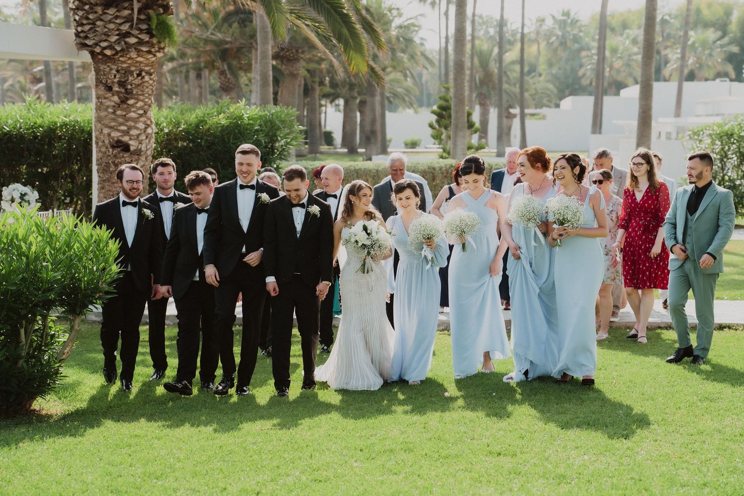 Wedding party poses together on green lawn, groomsmen in black tuxedos and bridesmaids in light blue gowns.