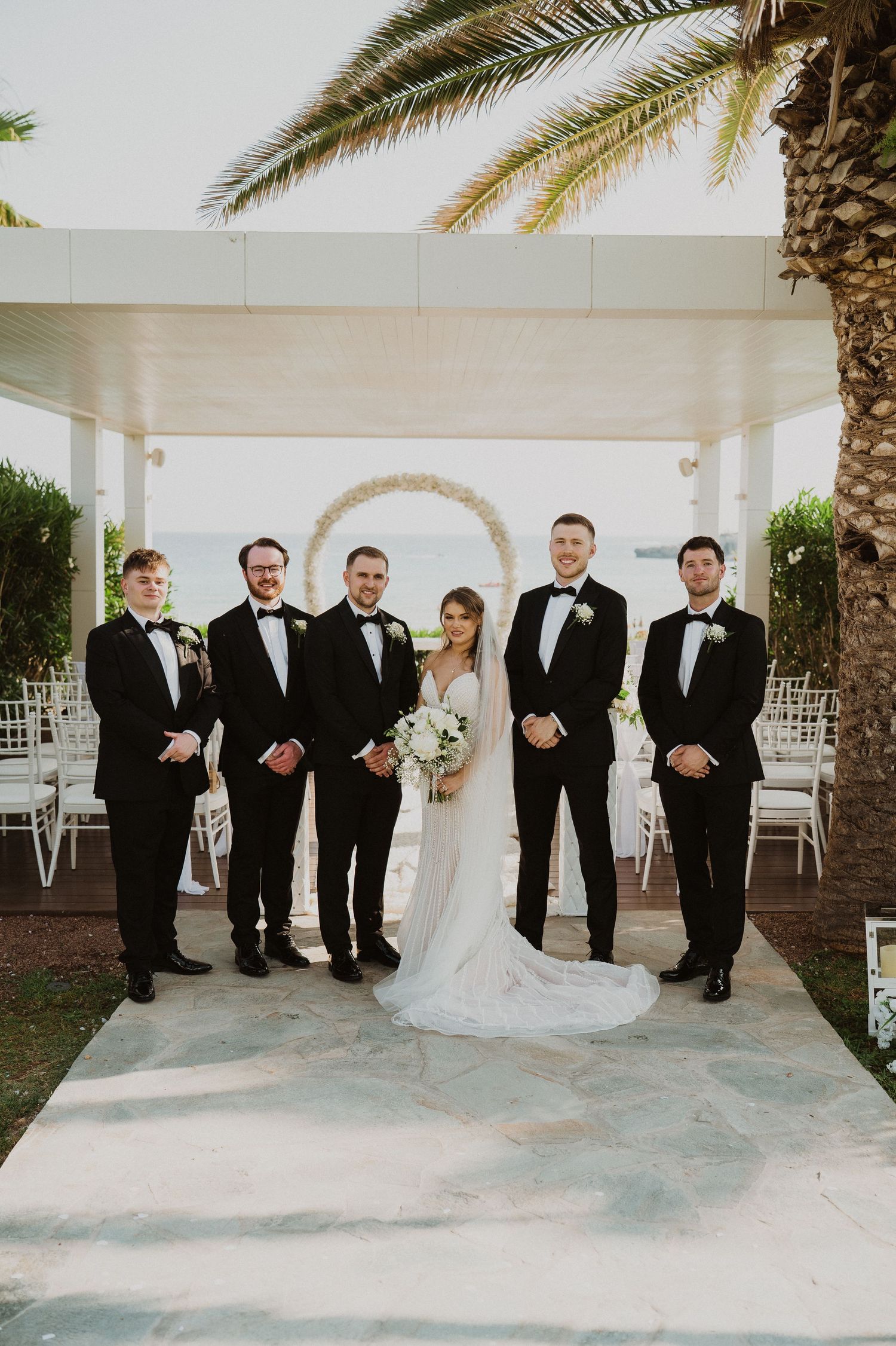 Bride and groom pose with groomsmen in black tuxedos at an outdoor wedding ceremony near a floral arch.