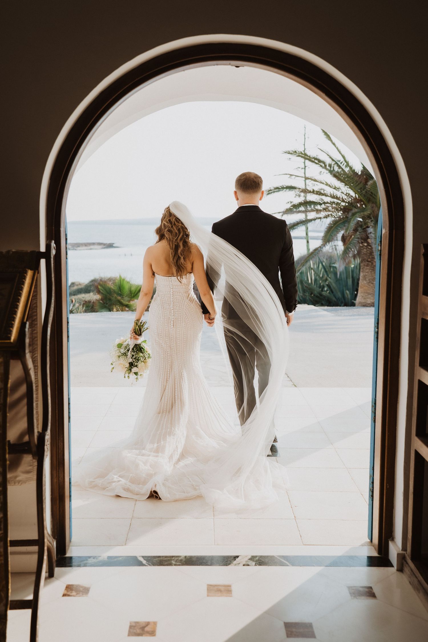 Newlyweds exit through ornate arched church doors toward ocean view, brides veil flowing in the breeze.