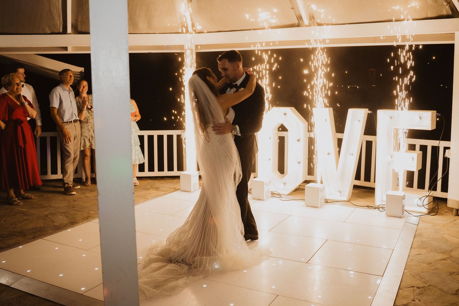 Bride and groom share first dance on illuminated floor with sparklers and large LOVE letters at outdoor wedding reception.