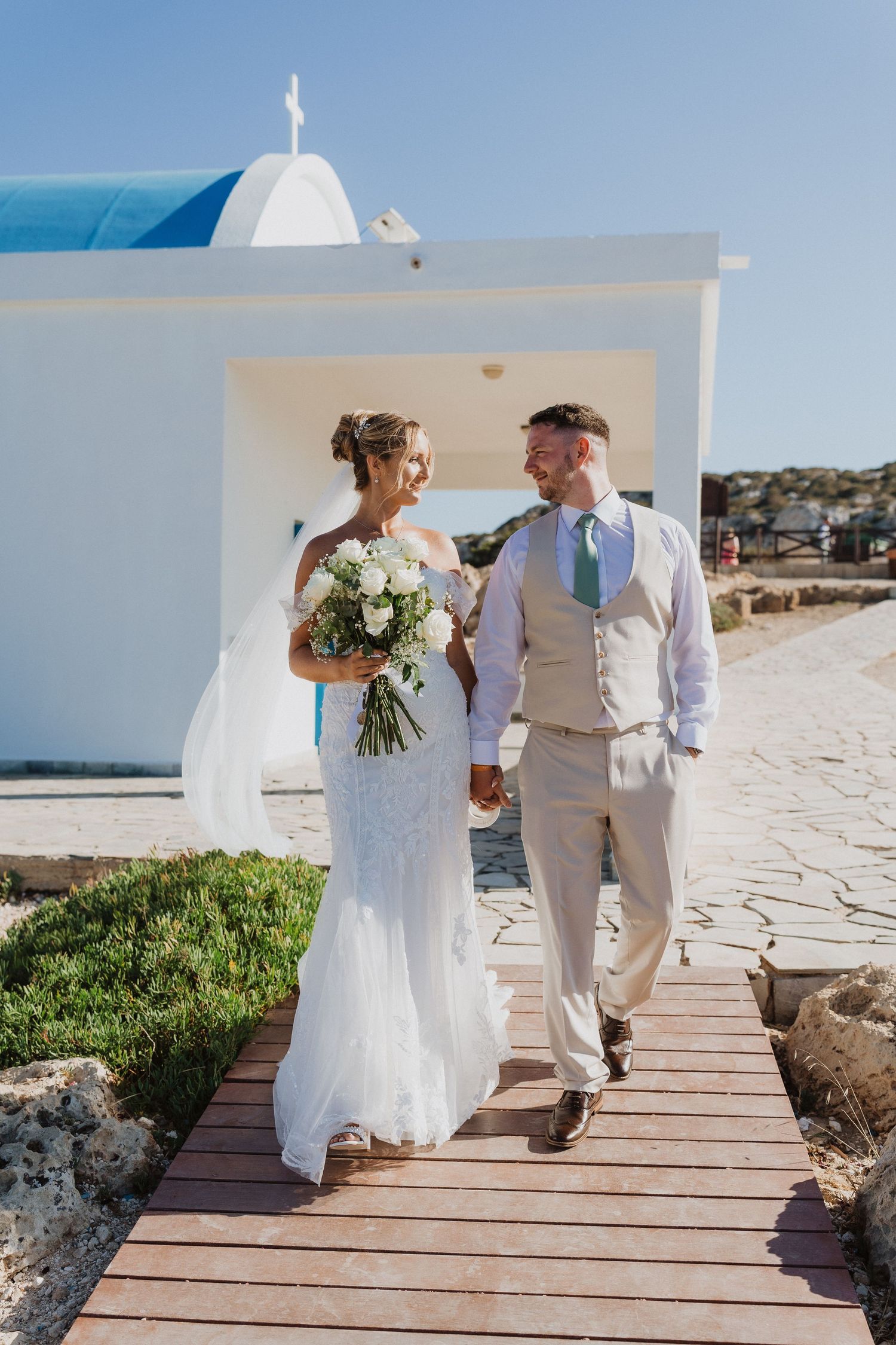 Couple in wedding attire walking along a wooden boardwalk on a sunny beach with white church dome in background.