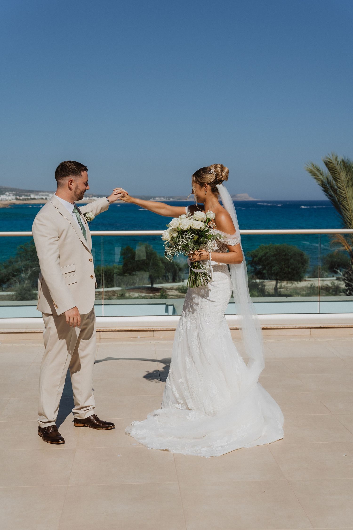 A couple in wedding attire dances playfully on a terrace overlooking a bright blue ocean on a sunny day.