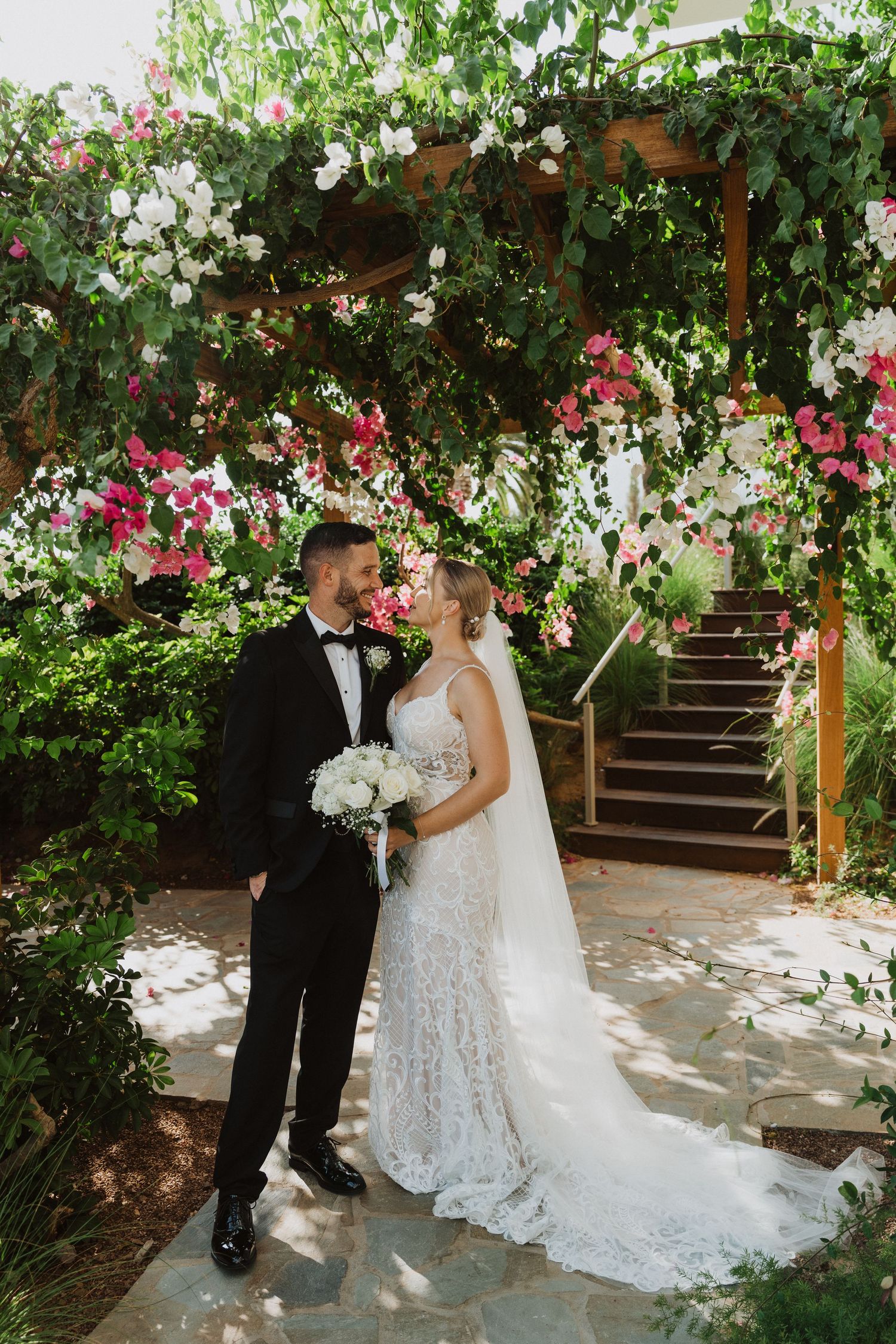 A couple shares a romantic moment under a wooden pergola covered in blooming white and pink flowers.