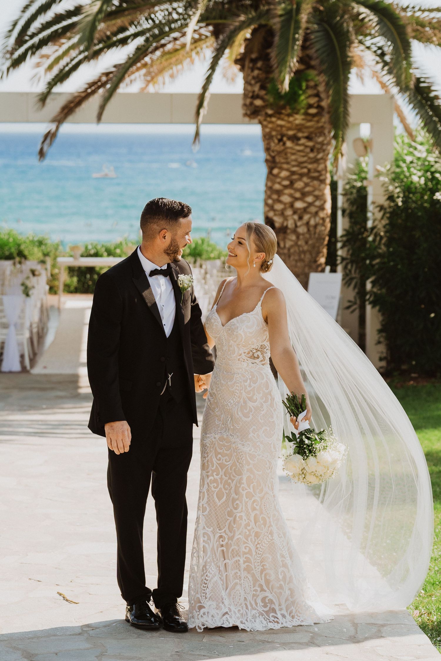 A bride and groom share a joyful moment under a palm tree by the ocean on their wedding day.
