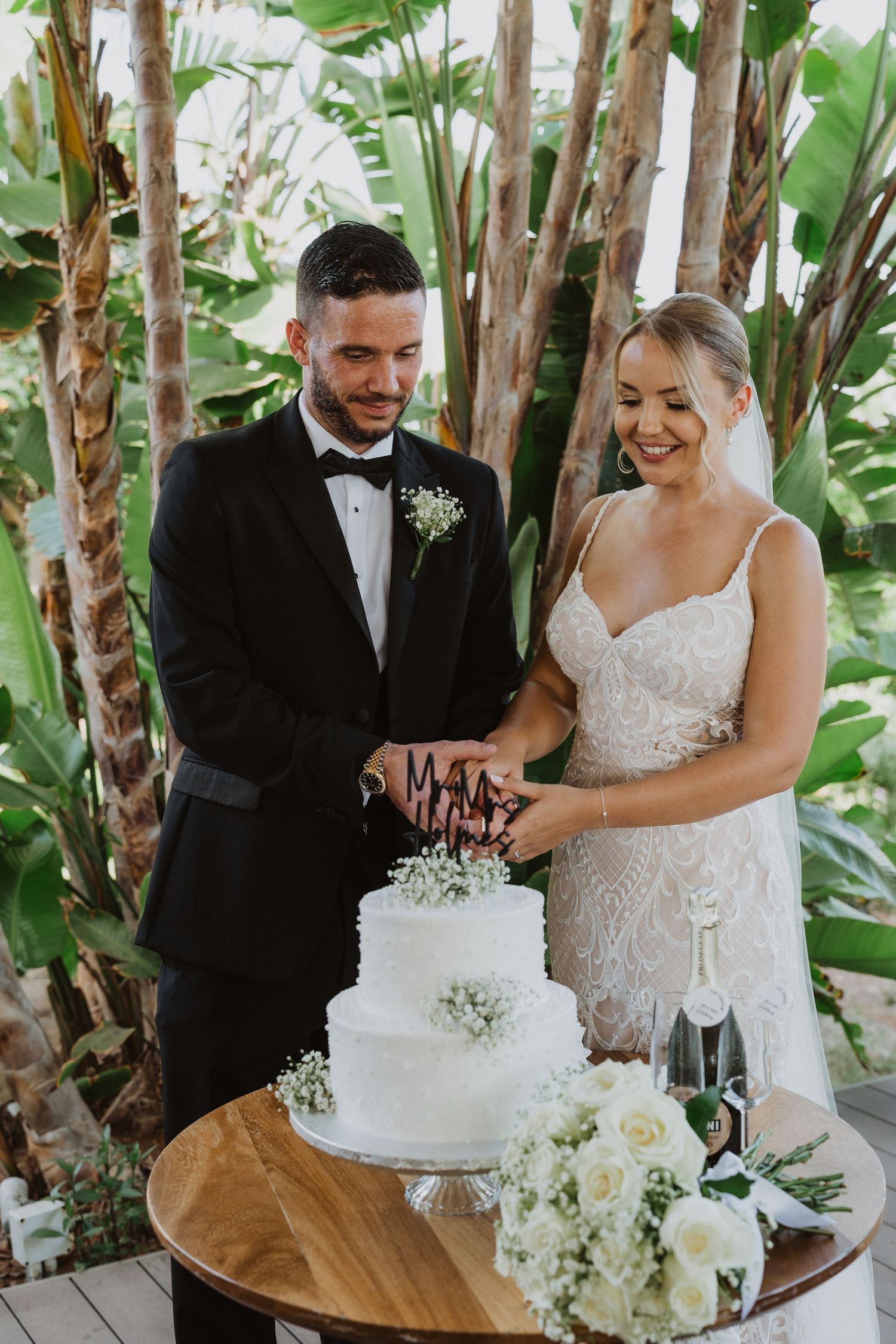 Wedding couple cutting their white tiered cake together during an outdoor tropical garden reception.