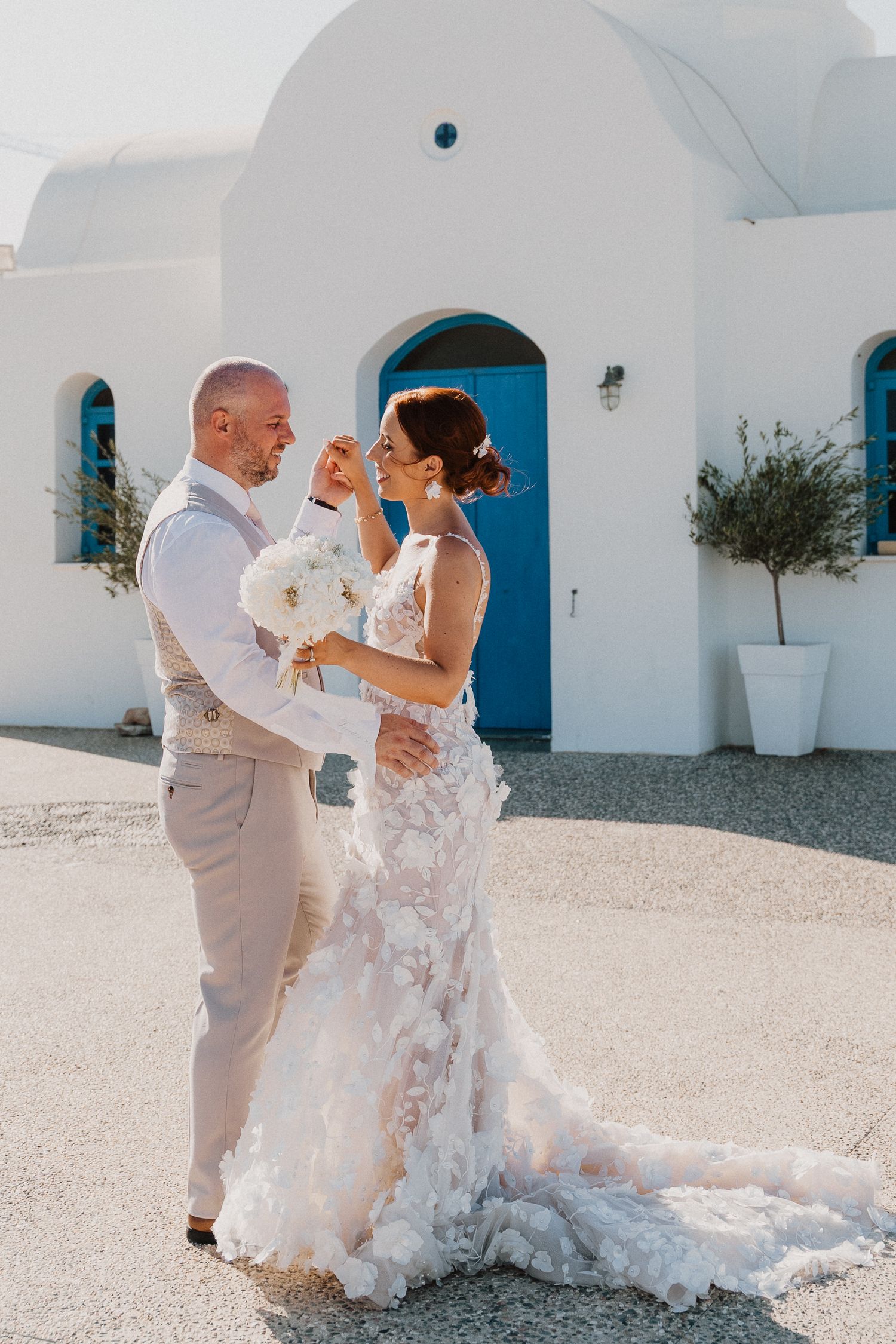 Happy couple shares a playful moment outside a white Mediterranean church in wedding attire.
