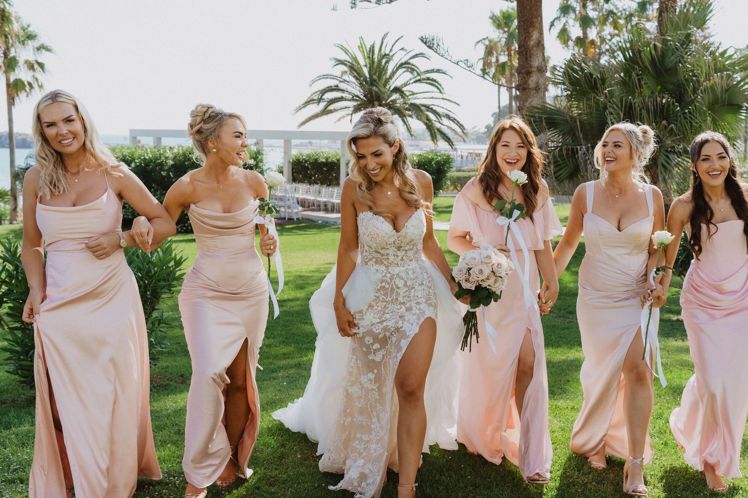 A wedding party wearing blush pink dresses poses on a sunny lawn with palm trees and ocean views in the background.