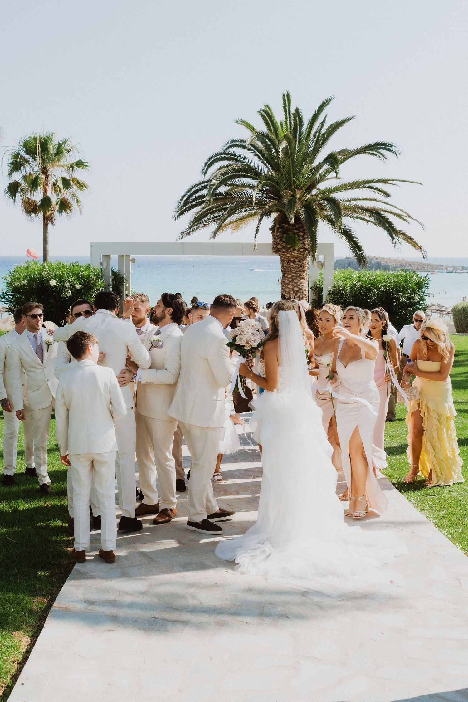 Wedding guests in white attire gather on a sunny terrace with palm trees during an elegant outdoor ceremony.
