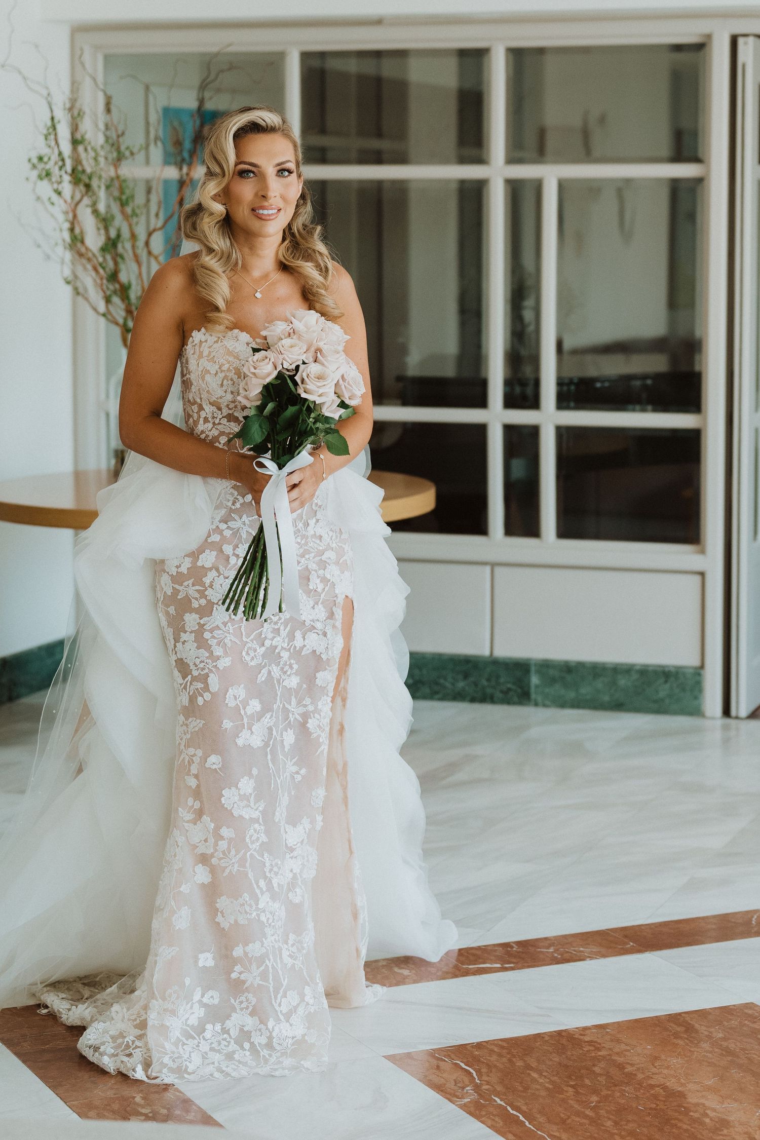A bride in a delicate white lace wedding gown with floral appliques holds a bouquet on a covered porch.