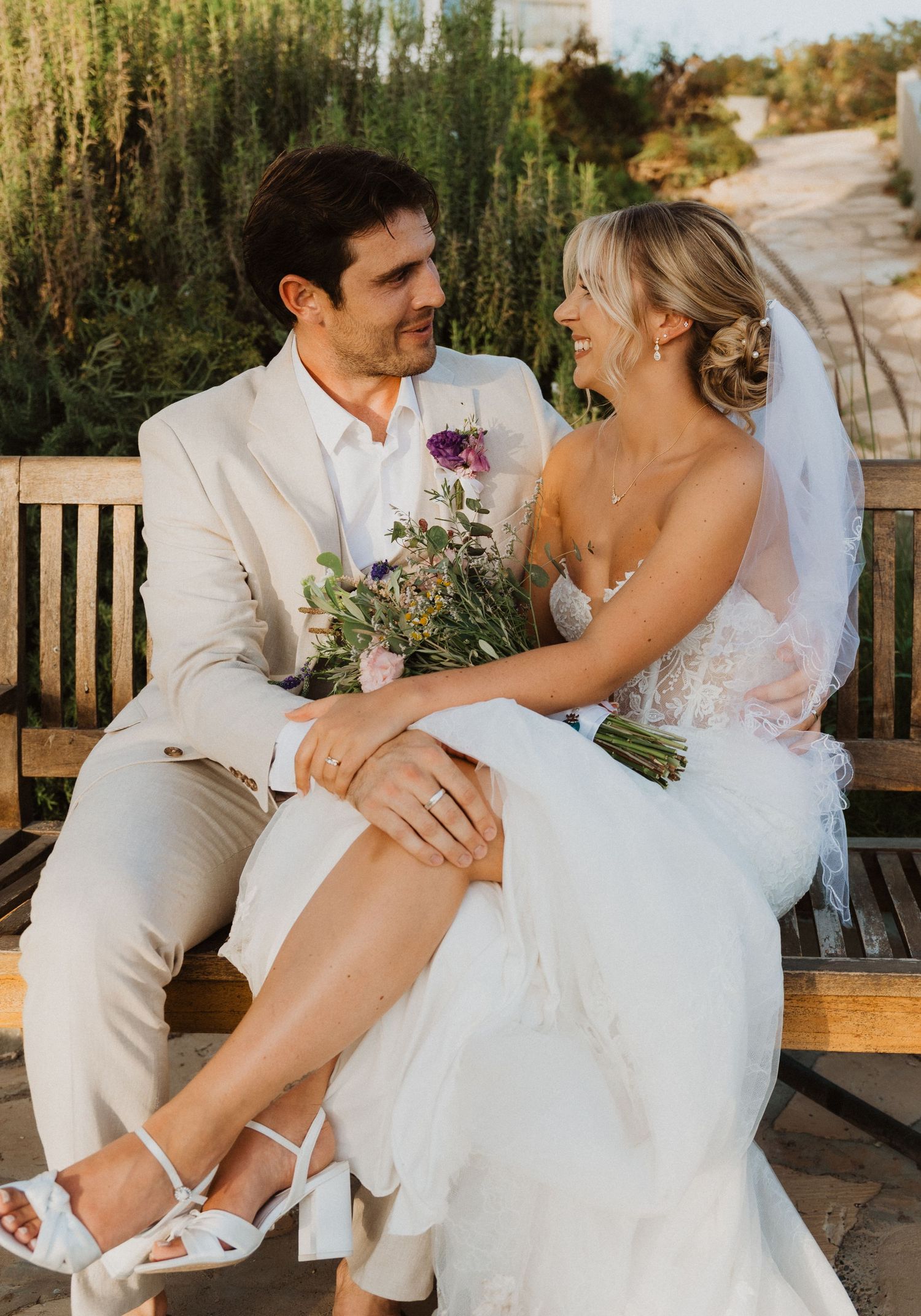 Bride and groom share intimate moments on wooden bench in lush garden setting.