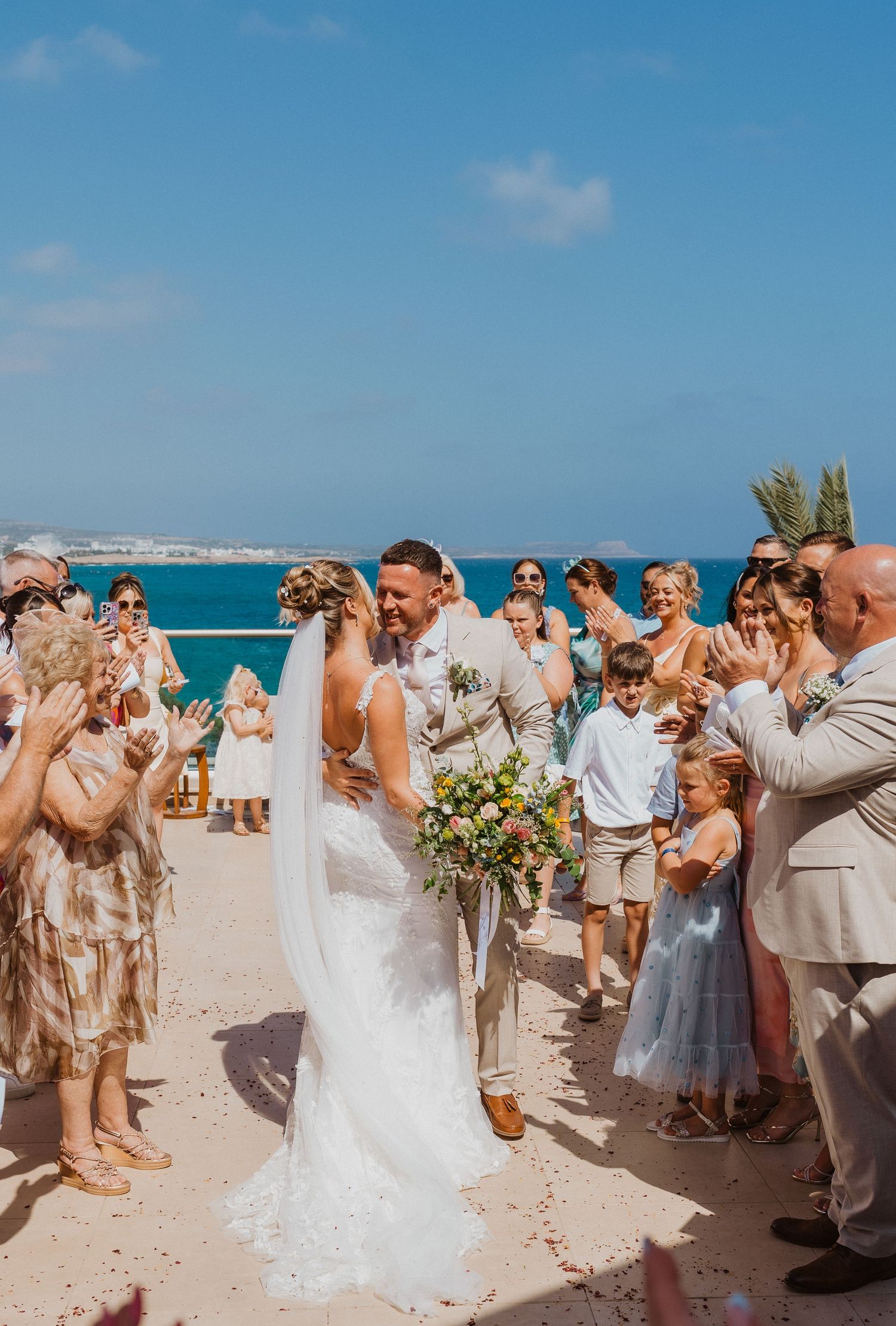 Wedding guests form a celebratory walkway on a sunny beach as newlyweds walk through with drinks raised in celebration.