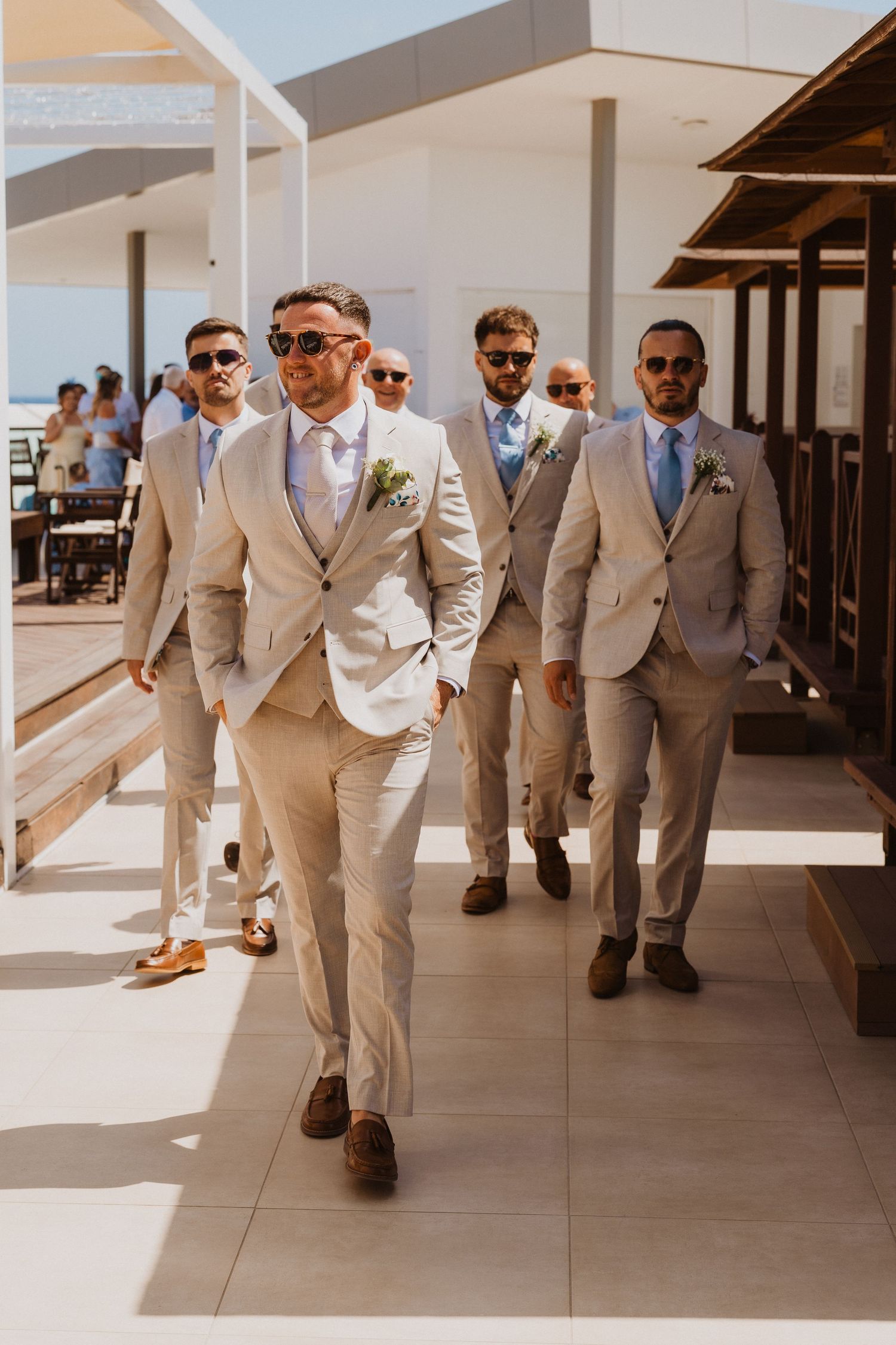 Groomsmen in beige suits and brown shoes walk confidently down a sunlit hallway in a modern building.