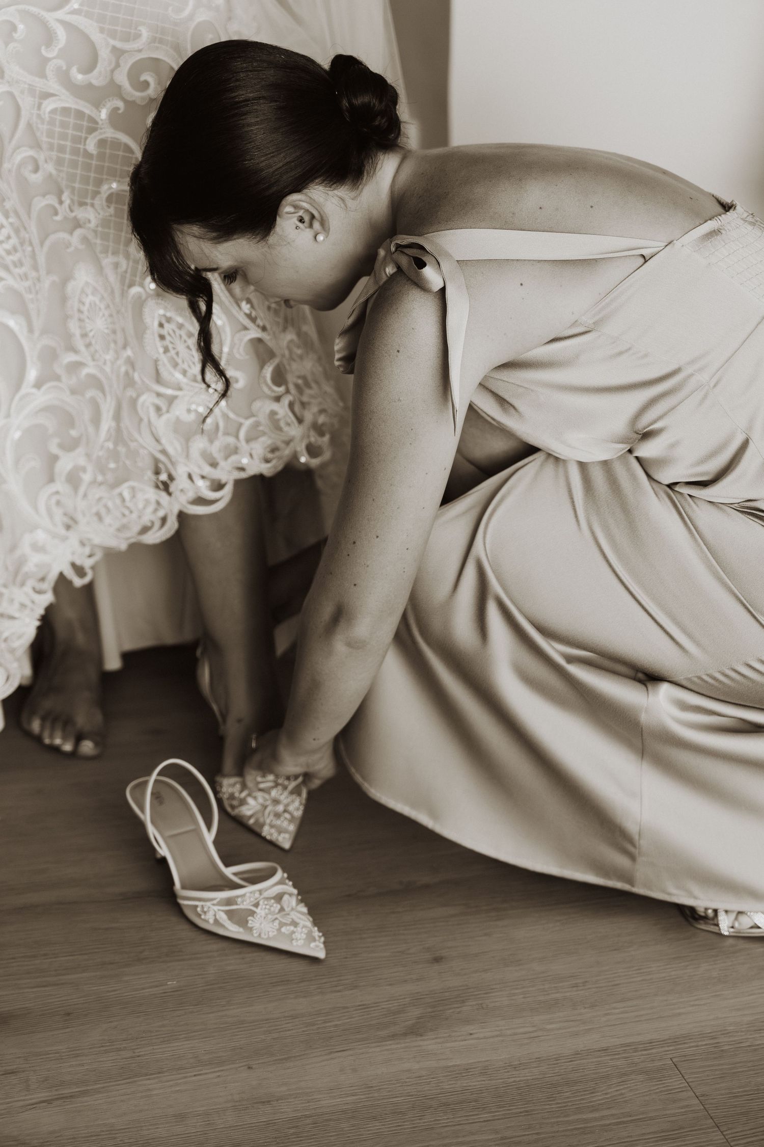 A person in a cream-colored dress reaches down to adjust an elegant pointed-toe wedding shoe on hardwood flooring.