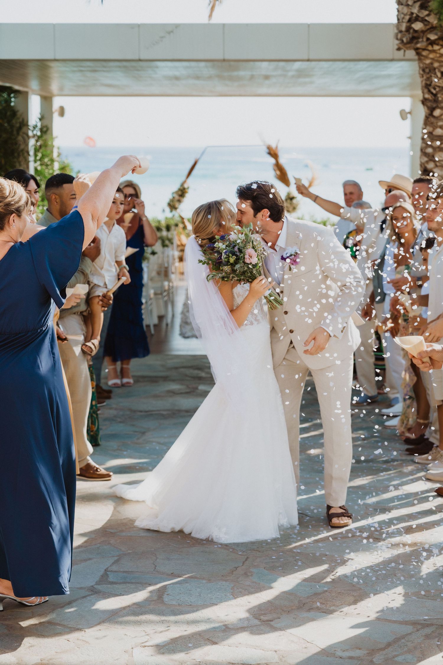 Wedding party celebrates with confetti toss under palm trees at outdoor venue.