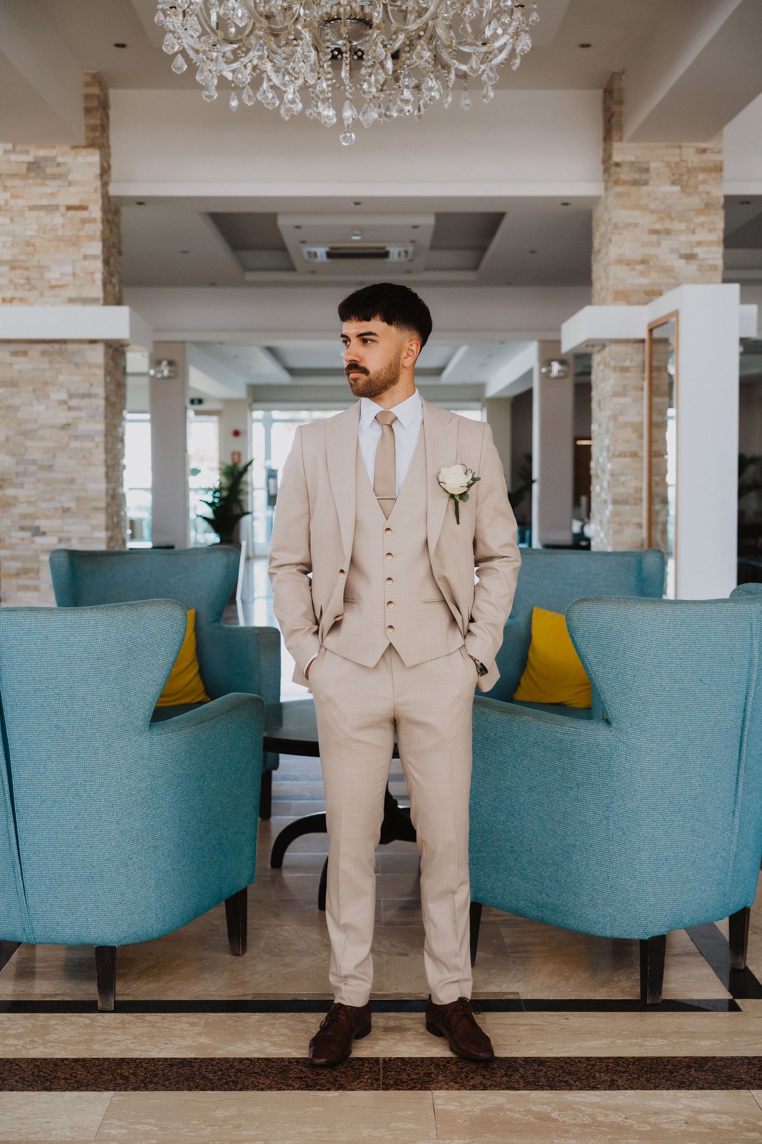A formal beige suit is showcased in a modern venue with blue upholstered seating.