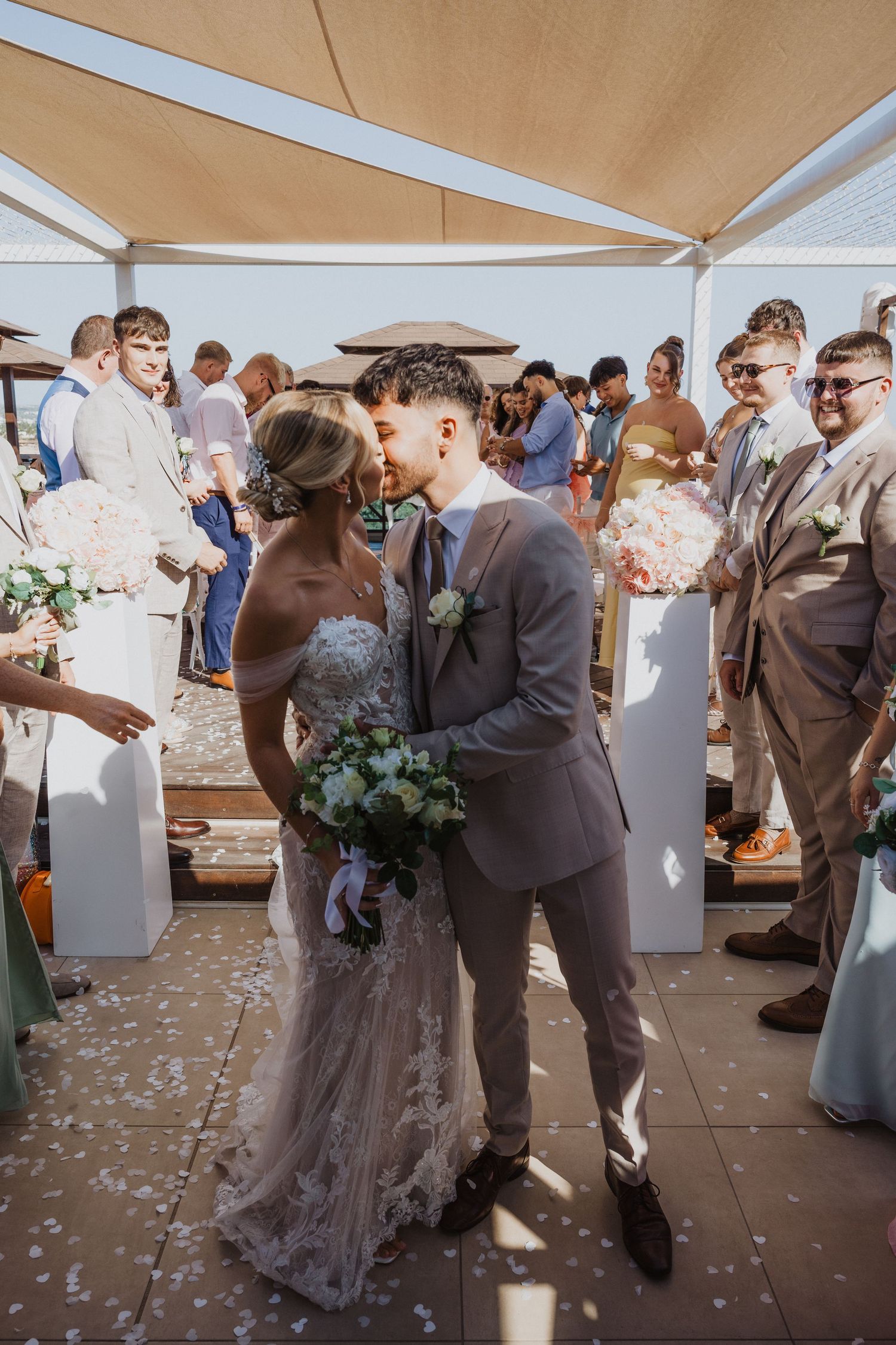 Romantic wedding moment captured under a canopy with guests and confetti in the background.