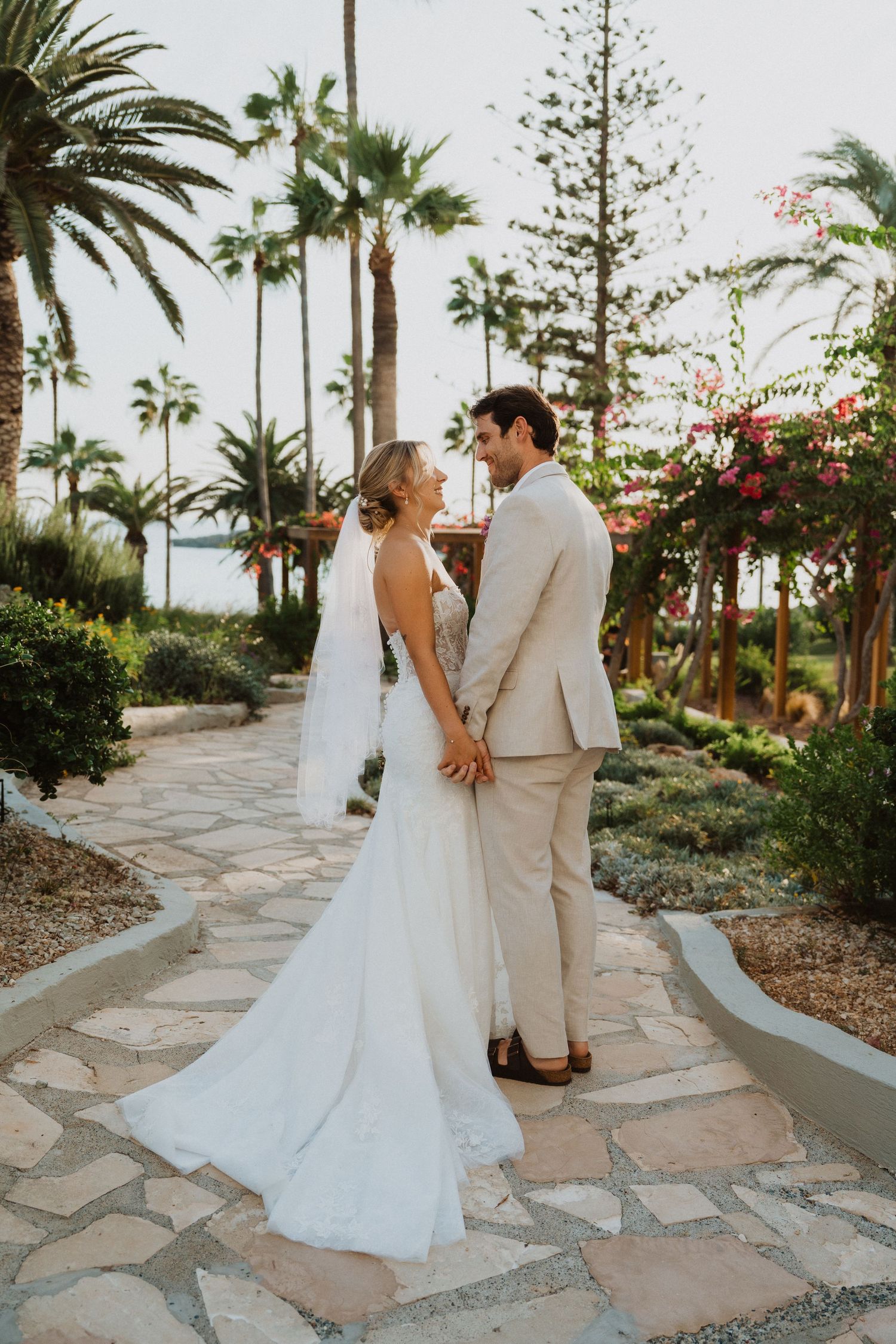 Newlyweds walk along a palm tree-lined stone pathway at their tropical venue.