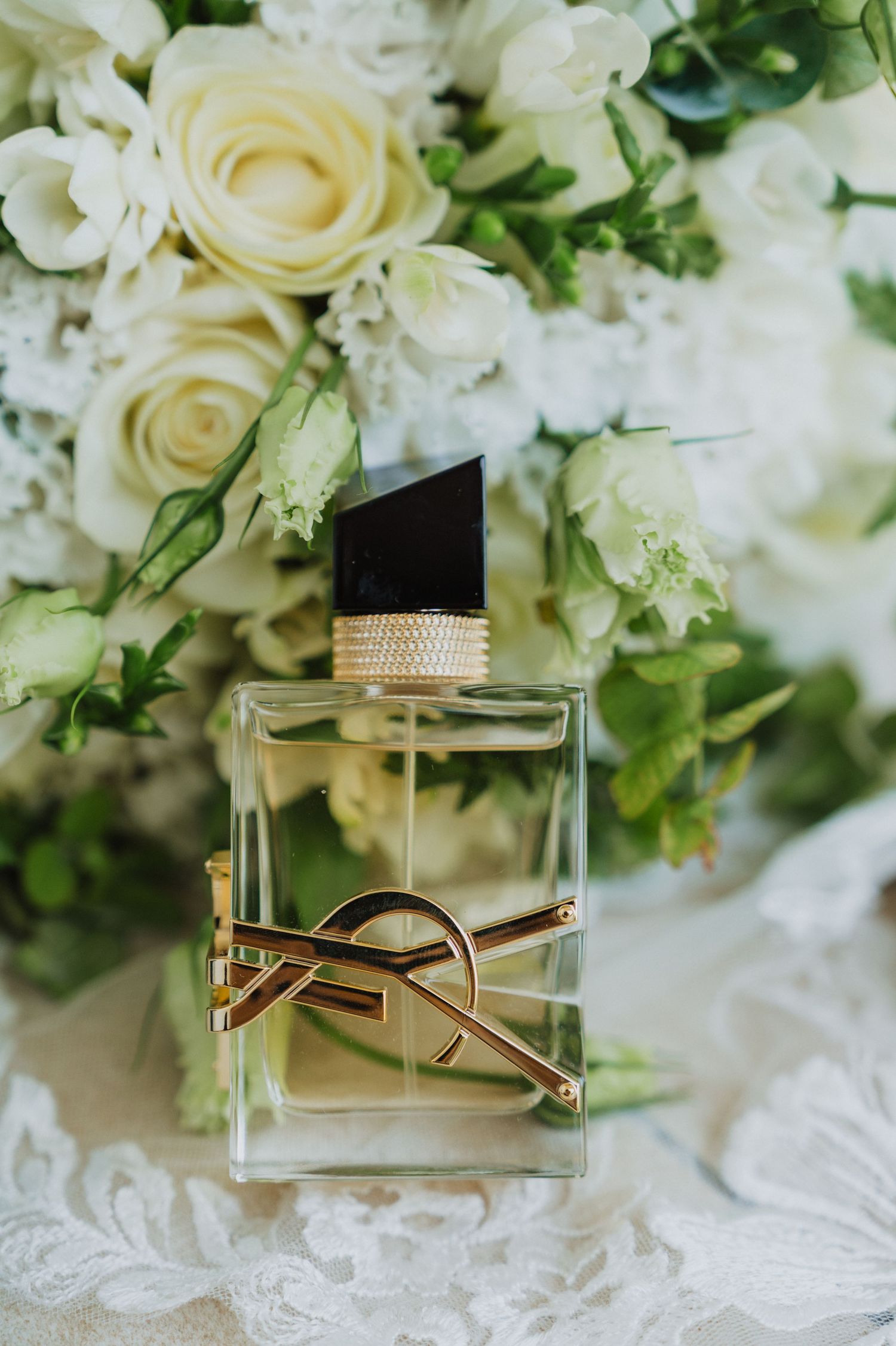 Elegant perfume bottle displayed among white roses and greenery on lace fabric.