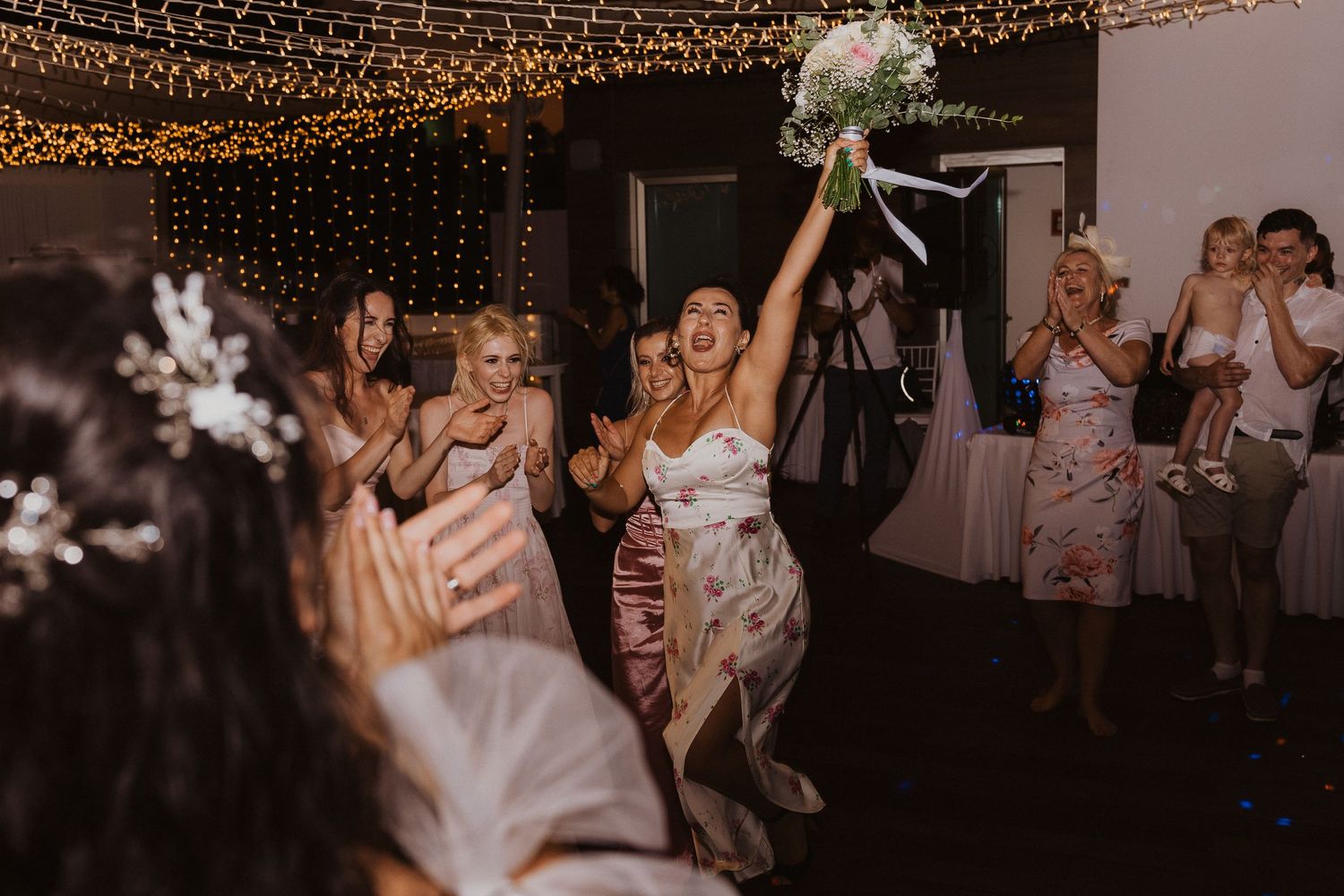 Wedding reception guests eagerly reach for the bouquet toss under romantic string light decor.