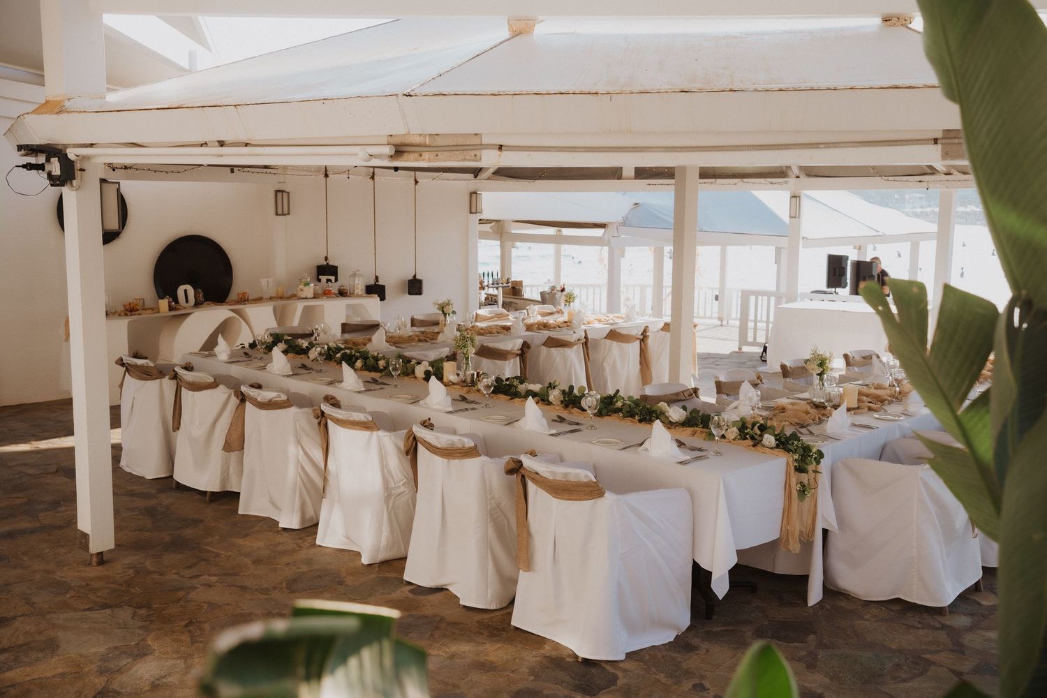 Elegant wedding reception table setup with white chair covers and floral centerpieces in a bright indoor venue.