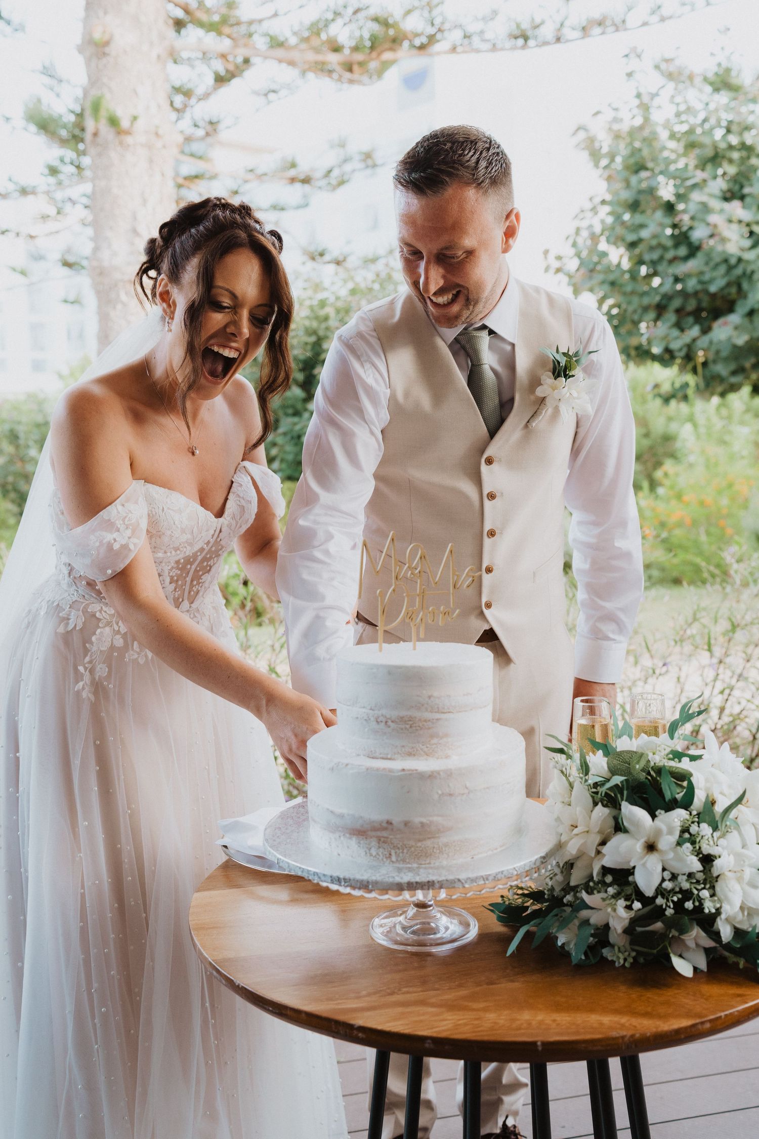 A couple shares a joyful moment while cutting their elegant white wedding cake at an outdoor reception.