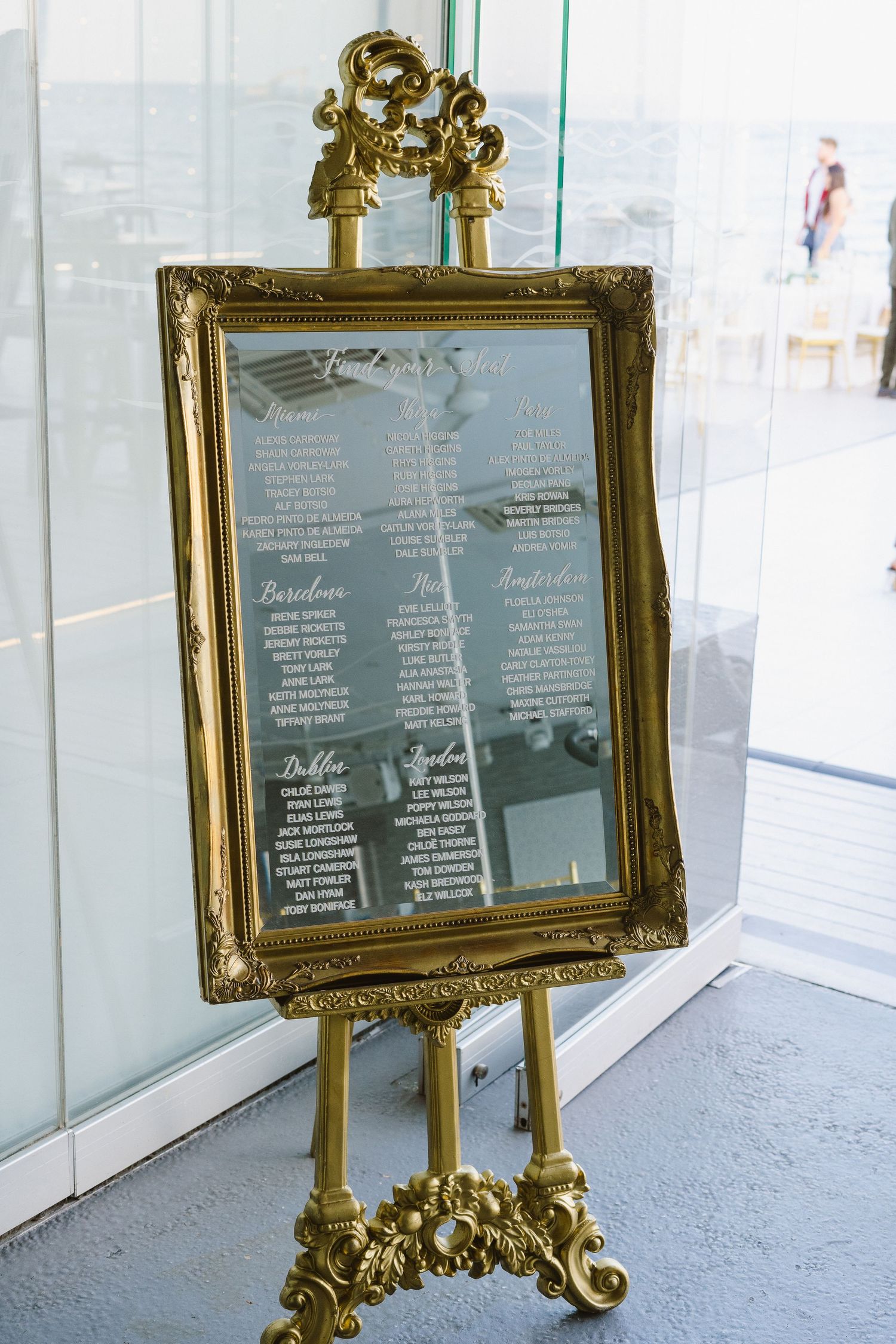 Ornate gold-framed mirror on decorative easel displaying elegant wedding seating assignments.