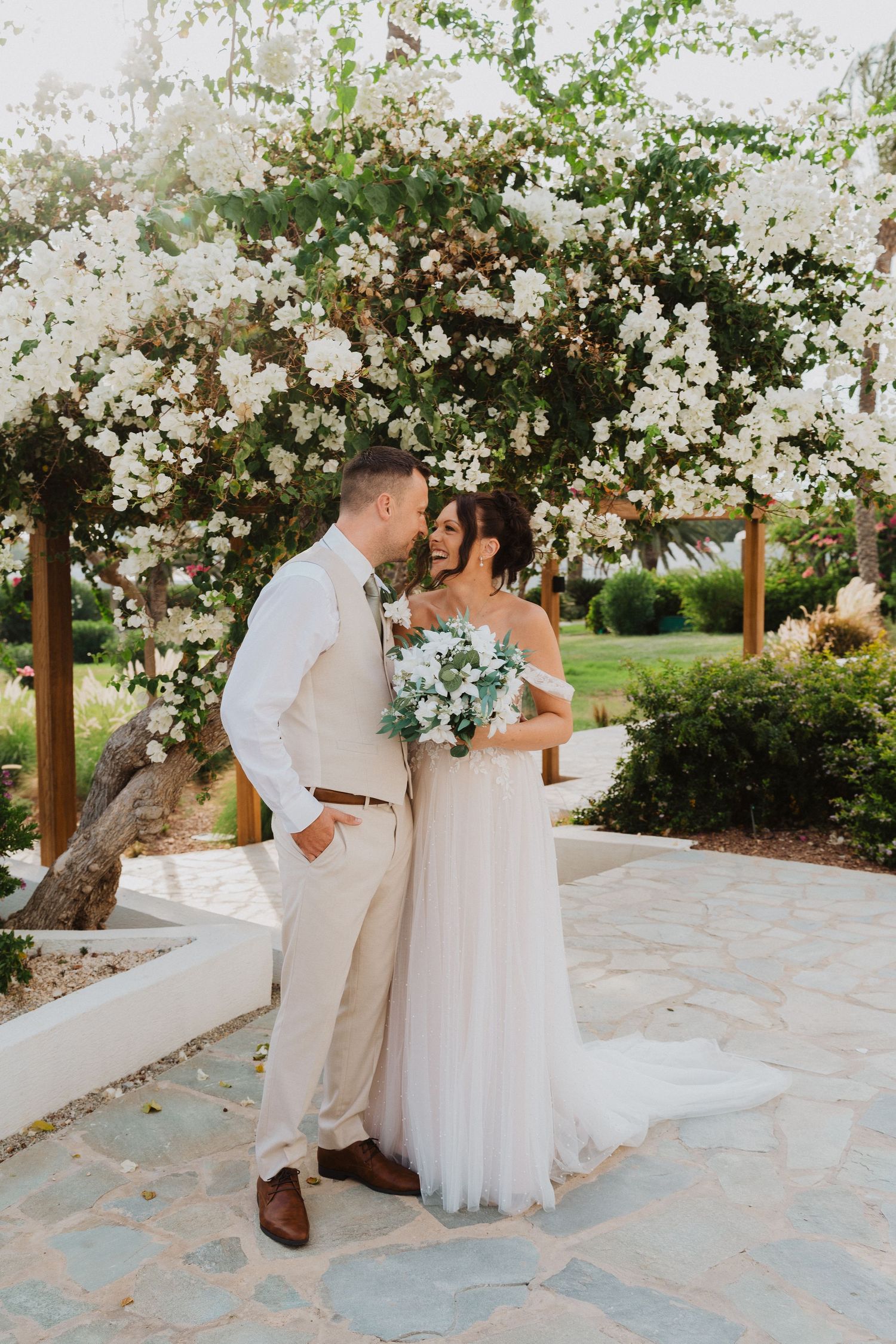 A romantic couple shares a tender moment under a white floral arch on a stone patio during their summer wedding celebration.
