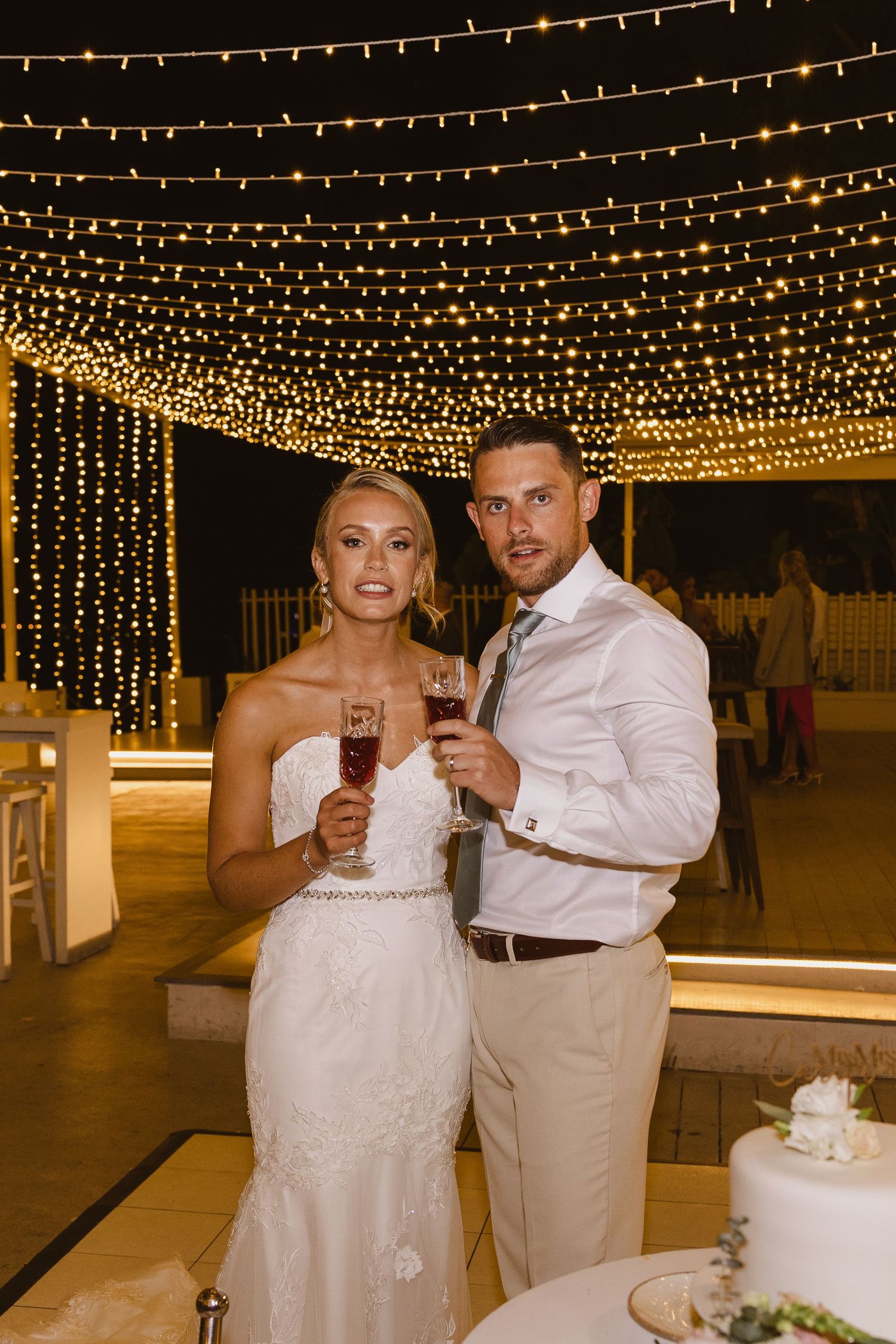 Newlyweds toast under a romantic canopy of twinkling string lights at an outdoor wedding reception.