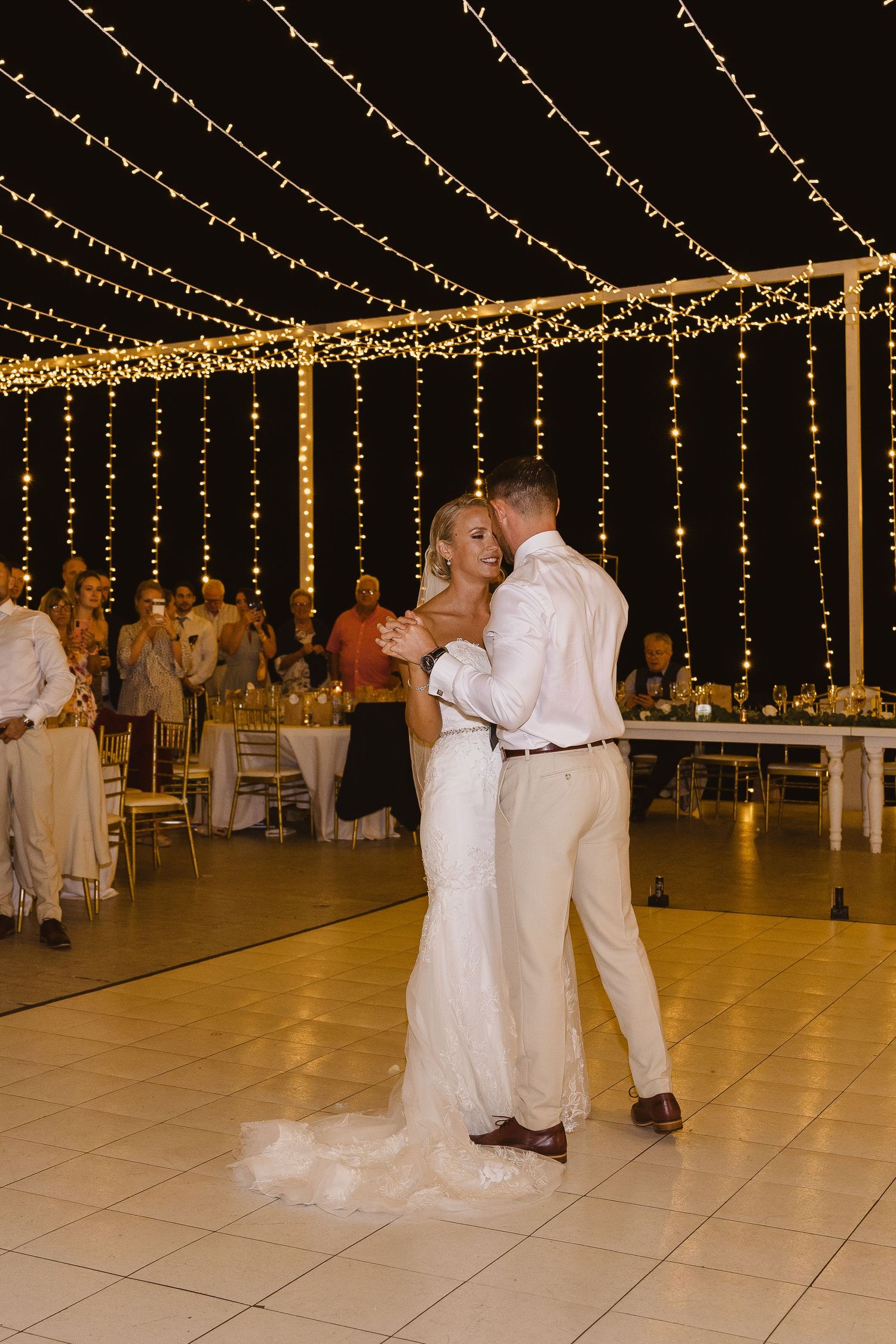 Newlyweds dance together under a canopy of warm fairy lights during their wedding reception.