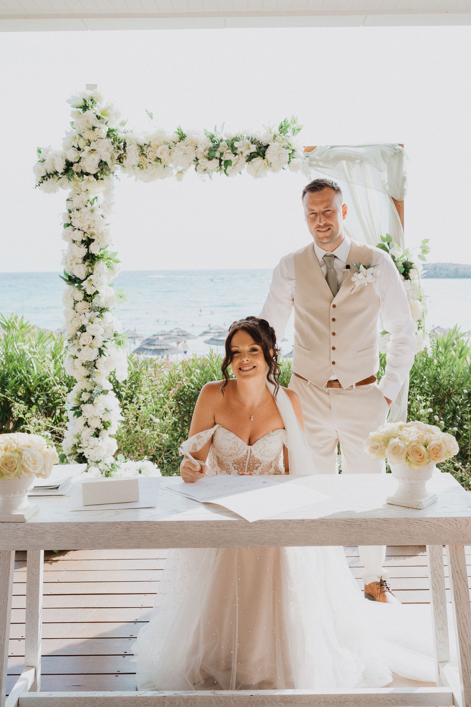 A couple sits at a white table under a floral arch during an outdoor beach wedding ceremony with ocean views.