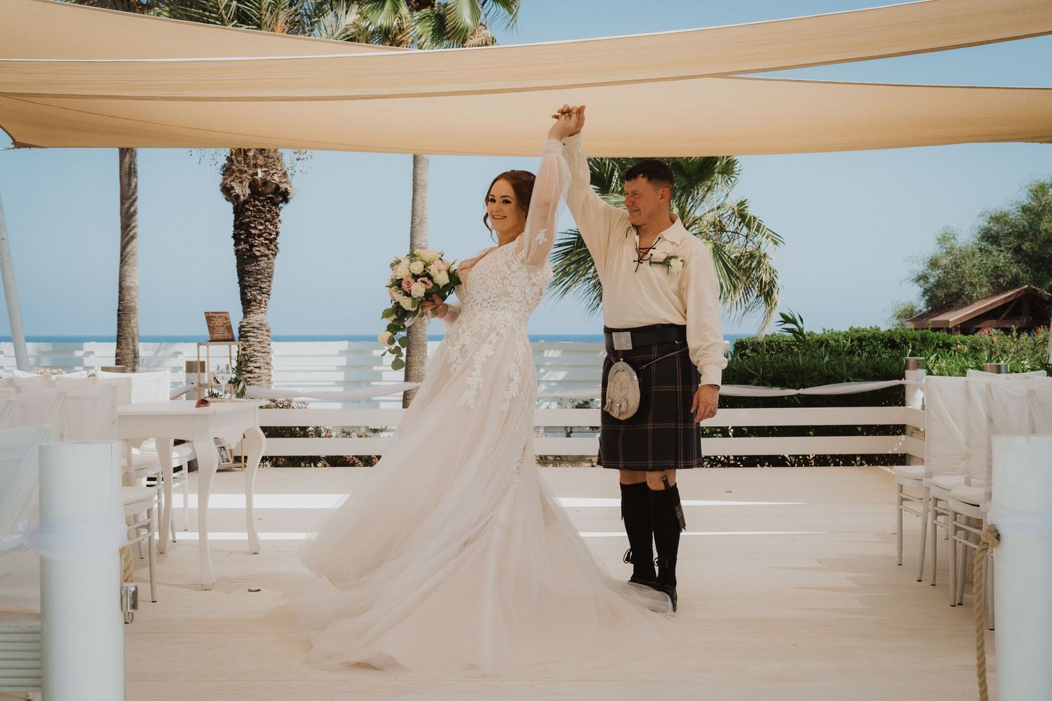 Newlyweds celebrate on outdoor terrace with ocean view, groom wearing traditional Scottish attire.