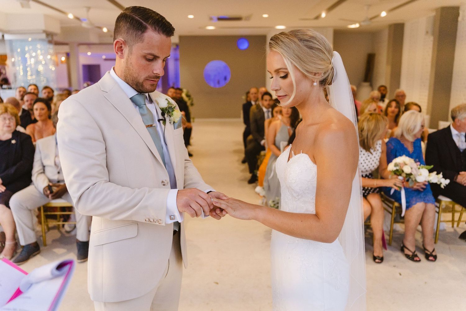 Exchange of rings during an indoor wedding ceremony with guests watching from the audience.