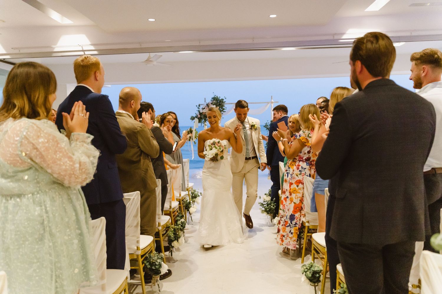 Wedding guests line the aisle as the couple makes their joyful exit in a bright indoor venue.