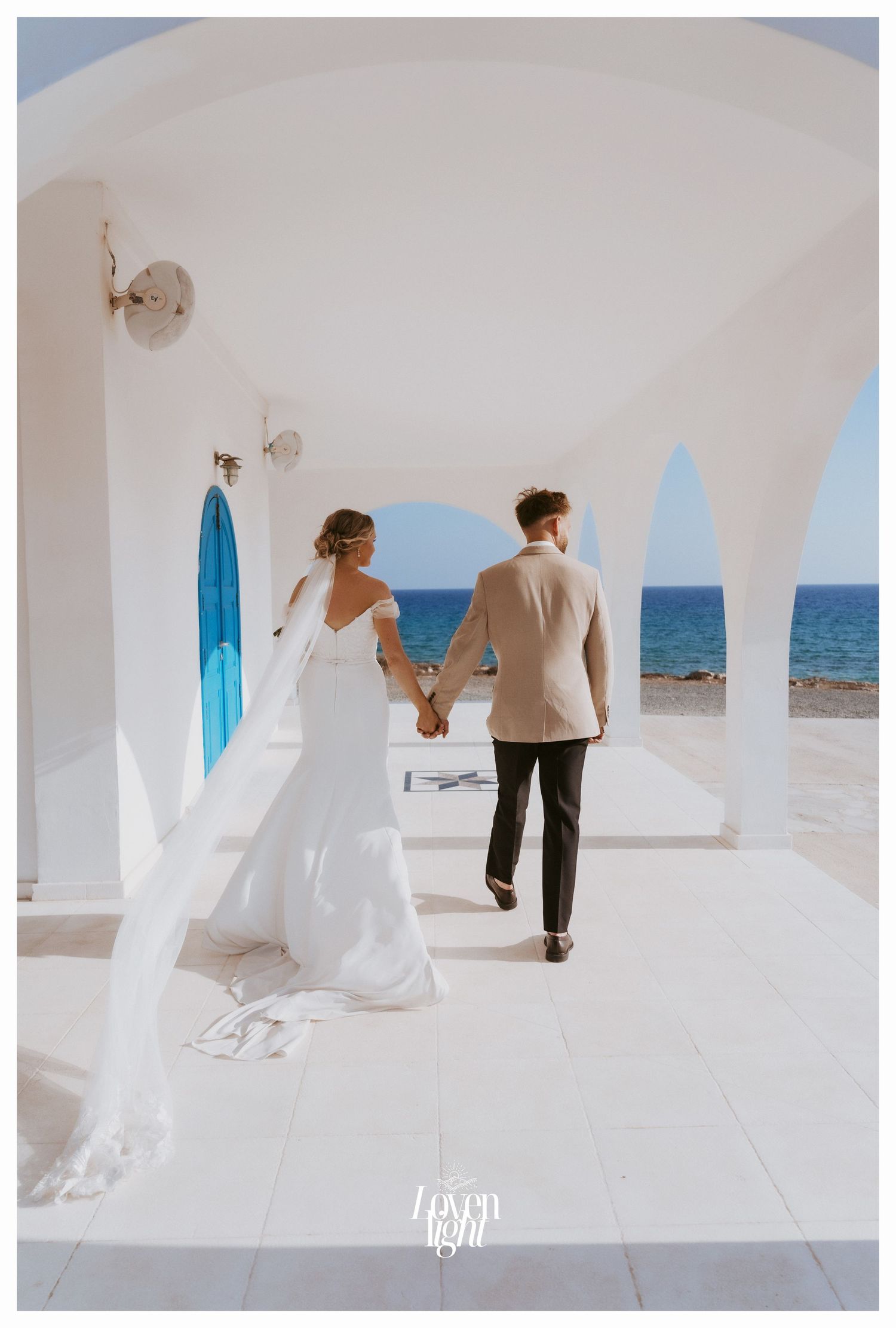 Newlyweds walking hand in hand through white Mediterranean archways overlooking ocean.