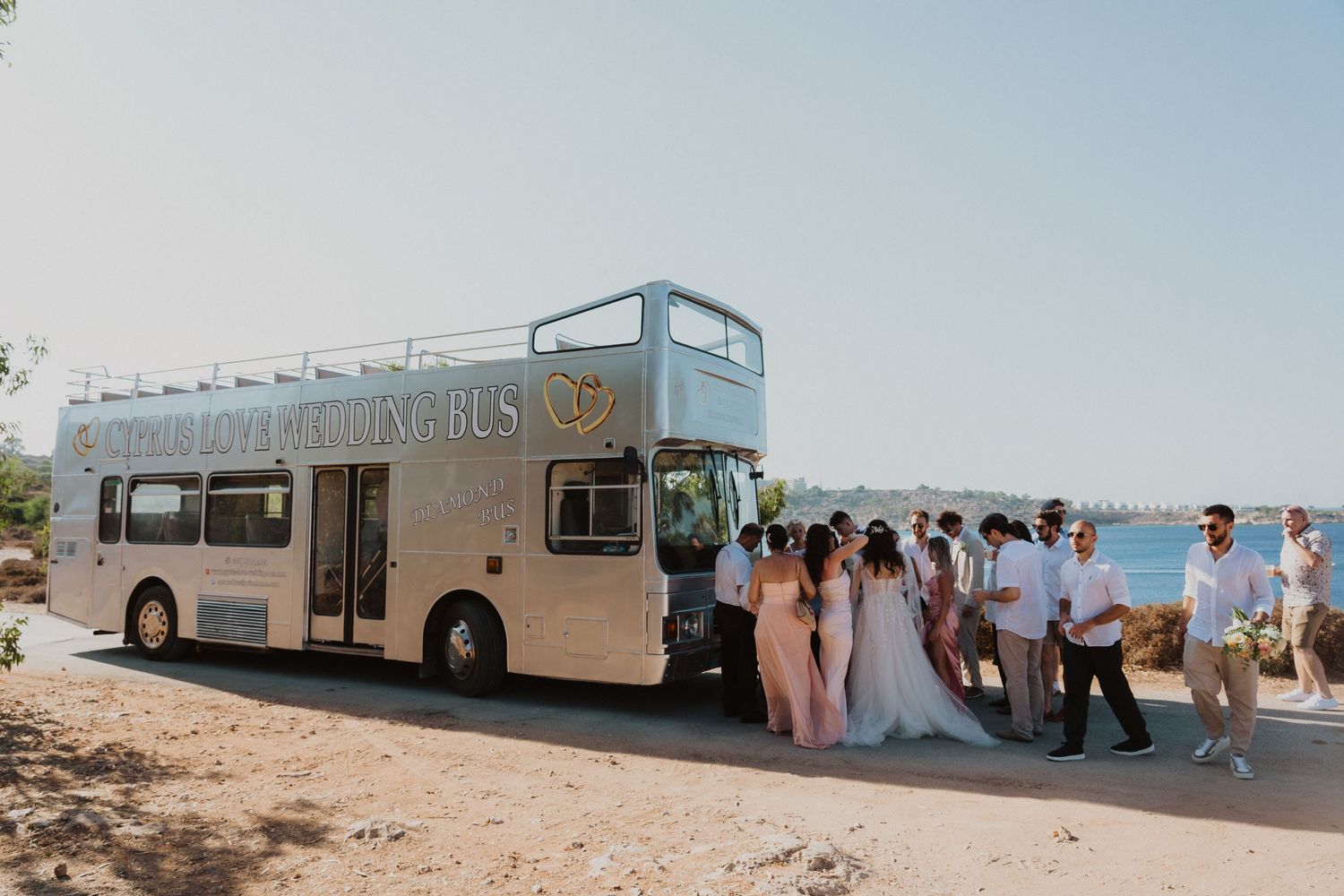 Light blue wedding bus parked by the coast with wedding party gathered around it.