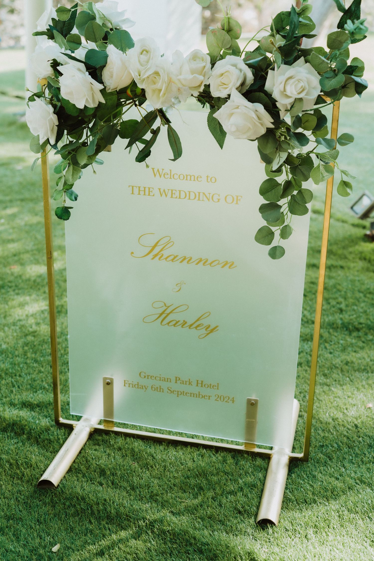 Wedding welcome sign with white roses and eucalyptus decorations in gold lettering on a clear acrylic stand.